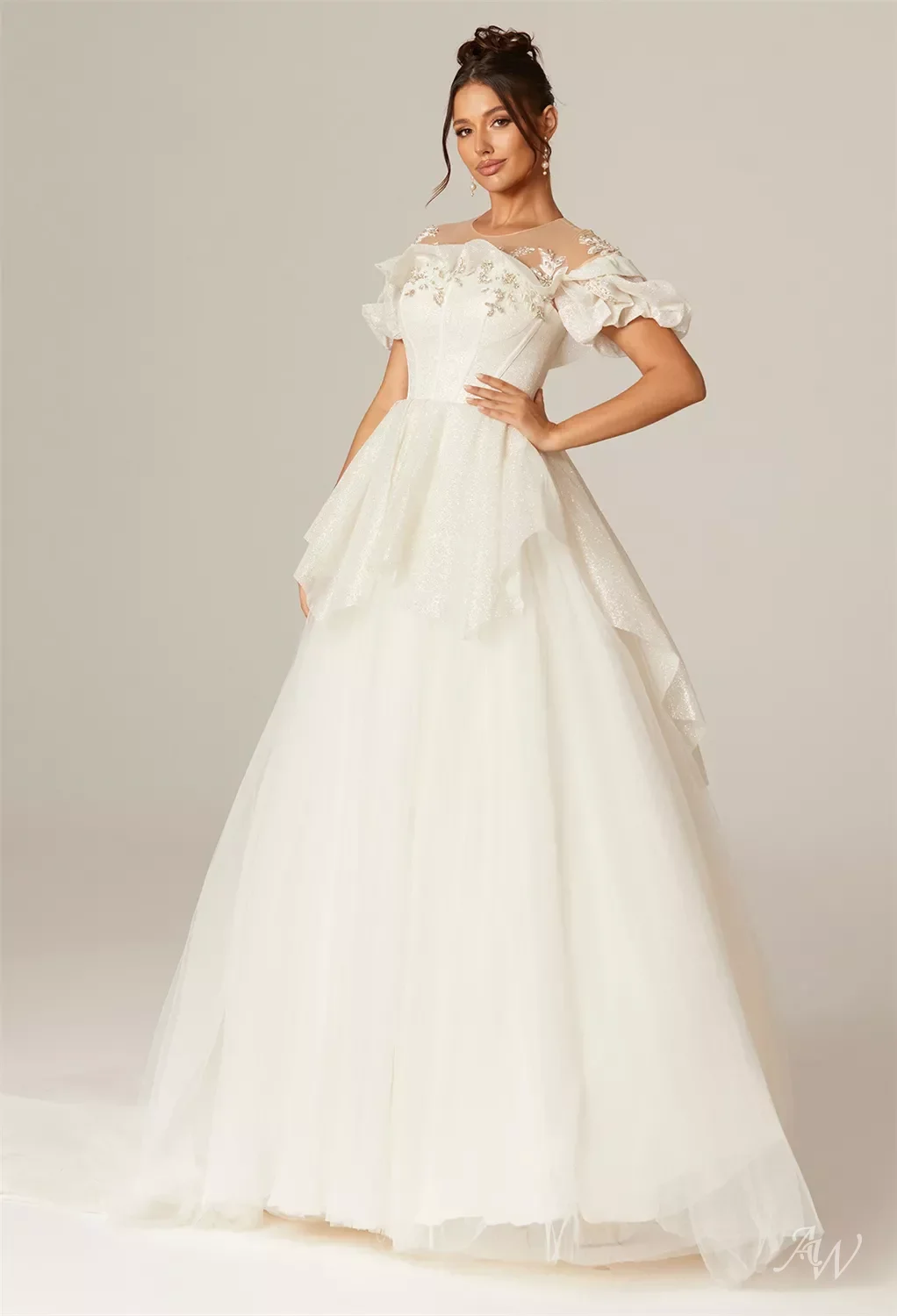 Woman in an elegant white wedding gown with lace and floral details, posing with one hand on her hip against a plain background.