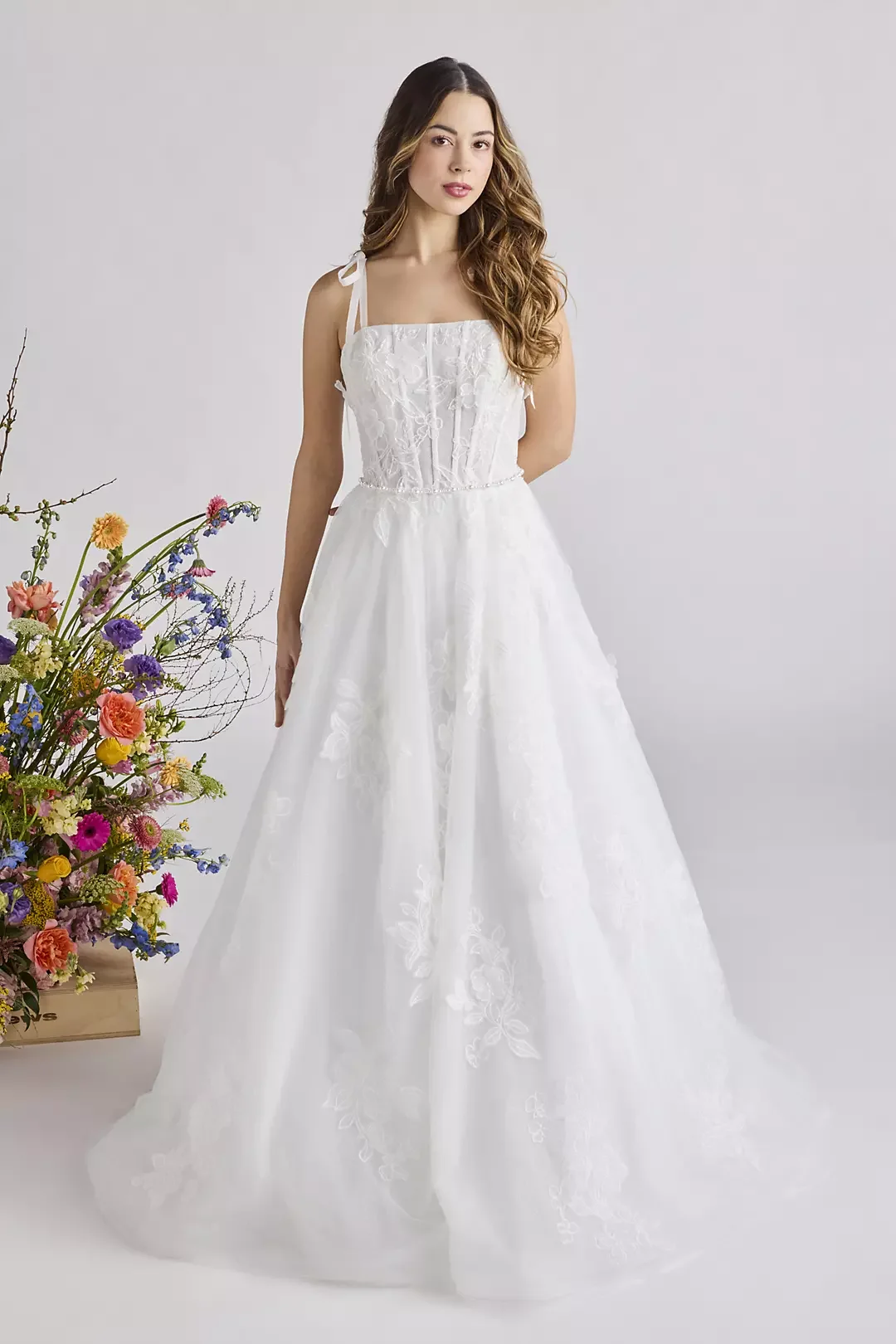 A woman wearing a white wedding dress with lace details, standing next to a colorful bouquet of flowers against a plain light background.