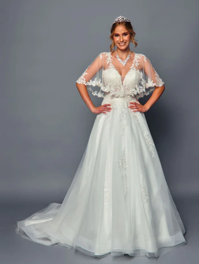 A woman wearing a white wedding gown with lace and embroidery, a sheer cape, a tiara, and a necklace, standing with hands on hips against a plain gray background.