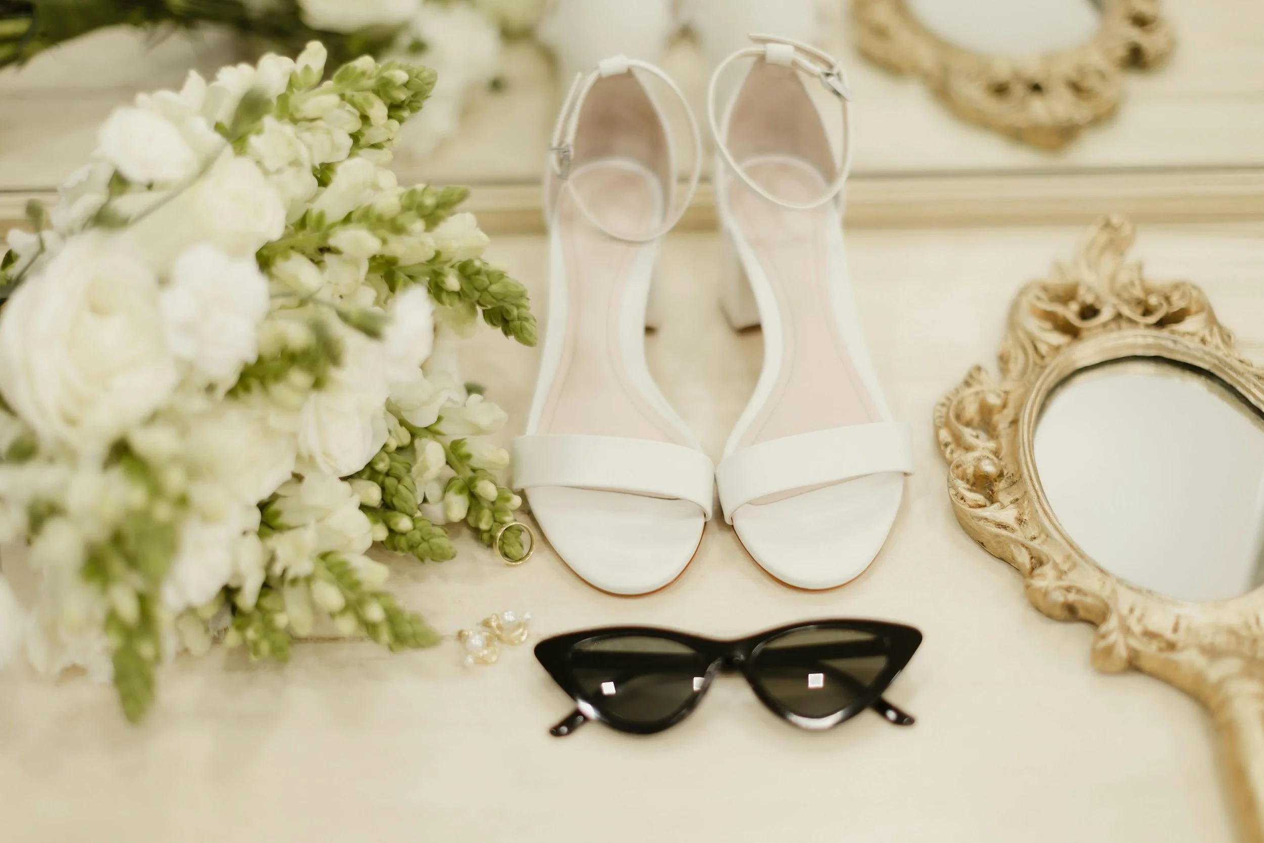 A pair of white wedding shoes, black sunglasses, a bouquet of white flowers, a small vintage mirror, and a pair of earrings arranged on a white surface.