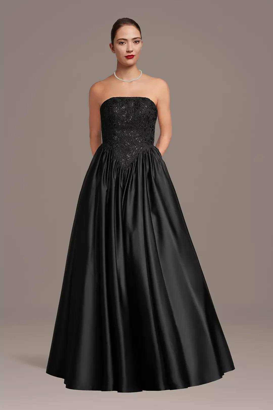 A woman wearing a strapless black evening gown with a lace and sequin bodice and full satin skirt, accessorized with a pearl necklace and red lipstick.