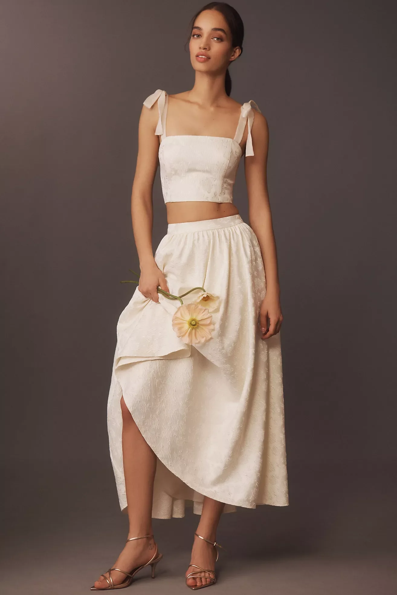 A woman standing against a plain dark background, wearing an elegant cream-colored, sleeveless crop top with ribbon shoulder straps, accessorized with high-heeled strappy sandals, and holding a peach-colored flower.