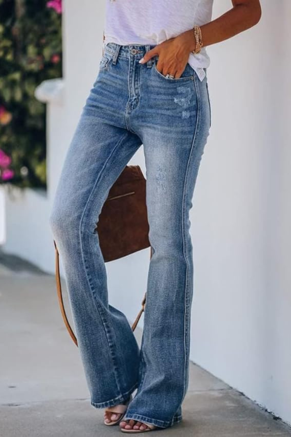 Person wearing high-waisted light blue flared jeans and a white top, with accessories and sandals, standing outdoors next to a white wall.