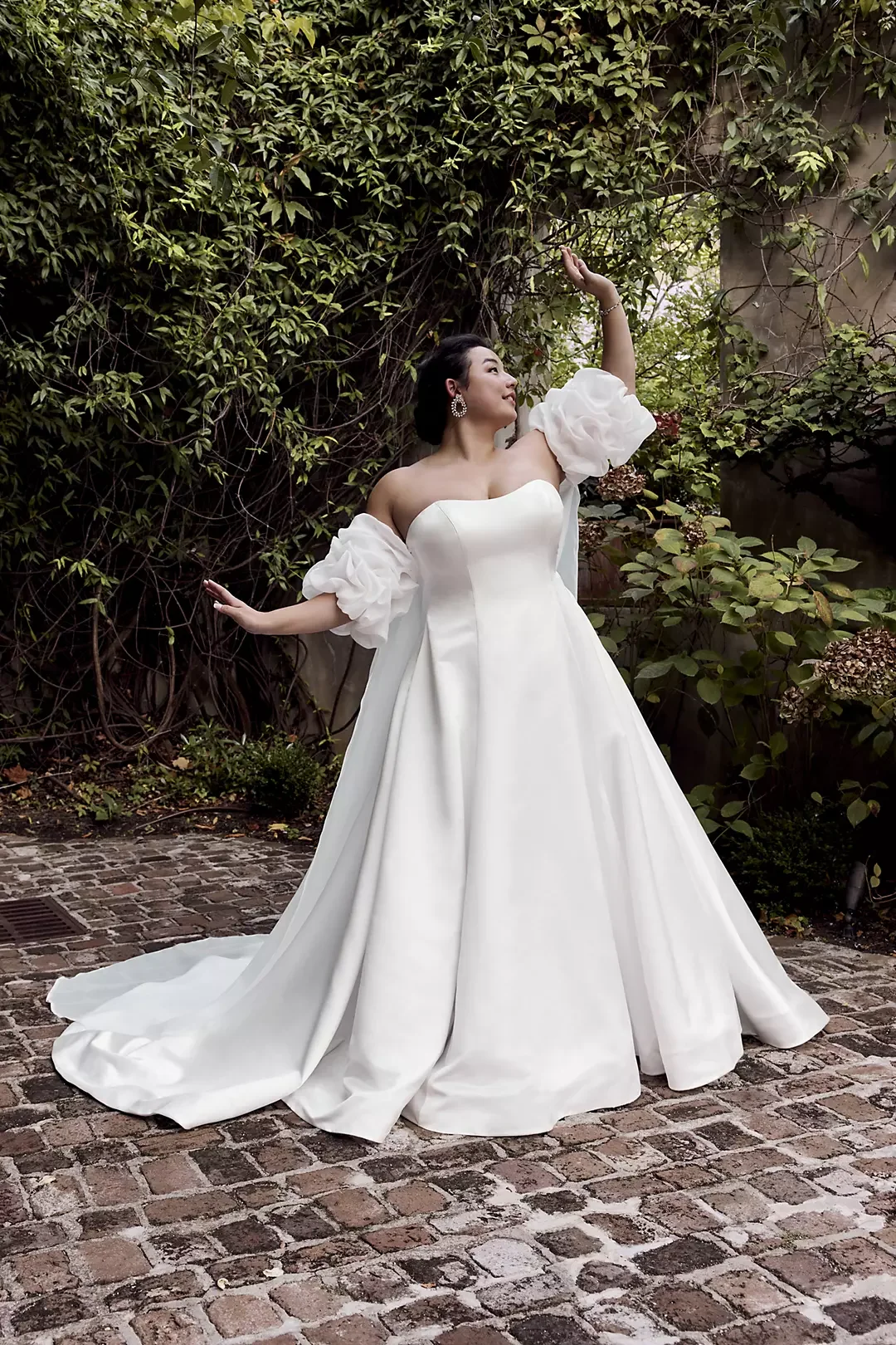 Woman in a white wedding gown with puffed sleeves, standing outdoors on a cobblestone path surrounded by greenery, posing gracefully with arms extended.
