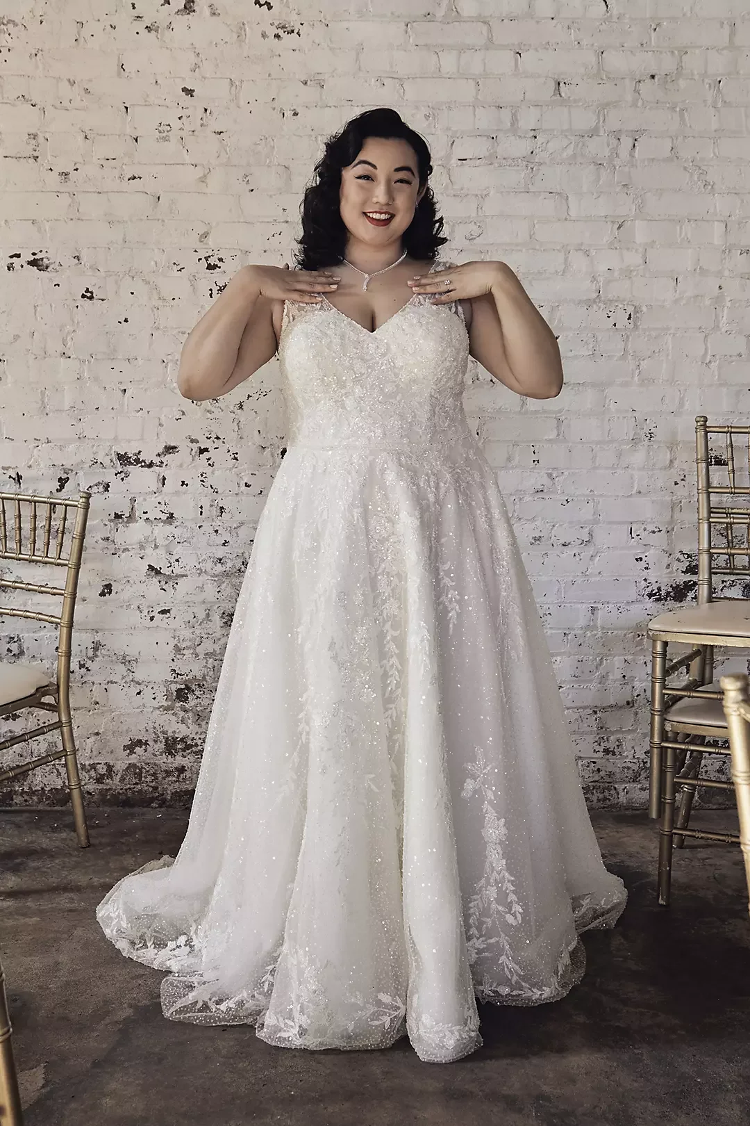 A woman in a white, sparkling wedding dress poses with her hands near her shoulders against a white brick wall background. There are gold chairs on either side of her.