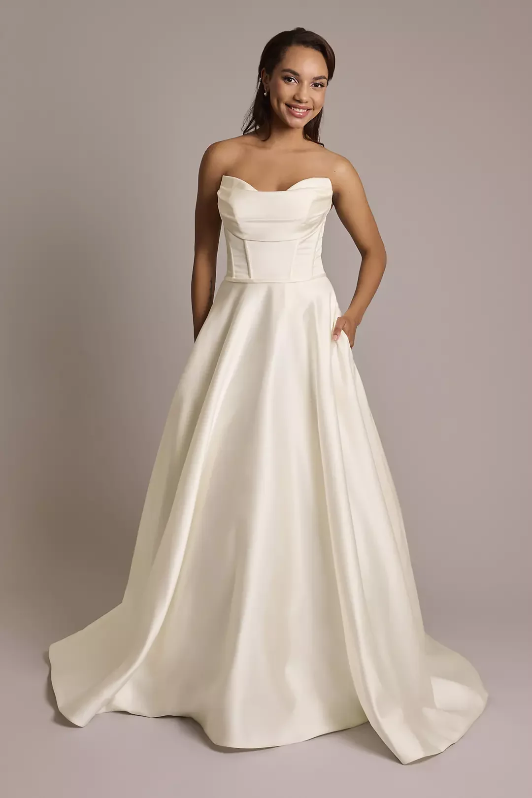 A woman wearing a white strapless wedding gown with a fitted bodice and full skirt, smiling and posing against a plain neutral background.