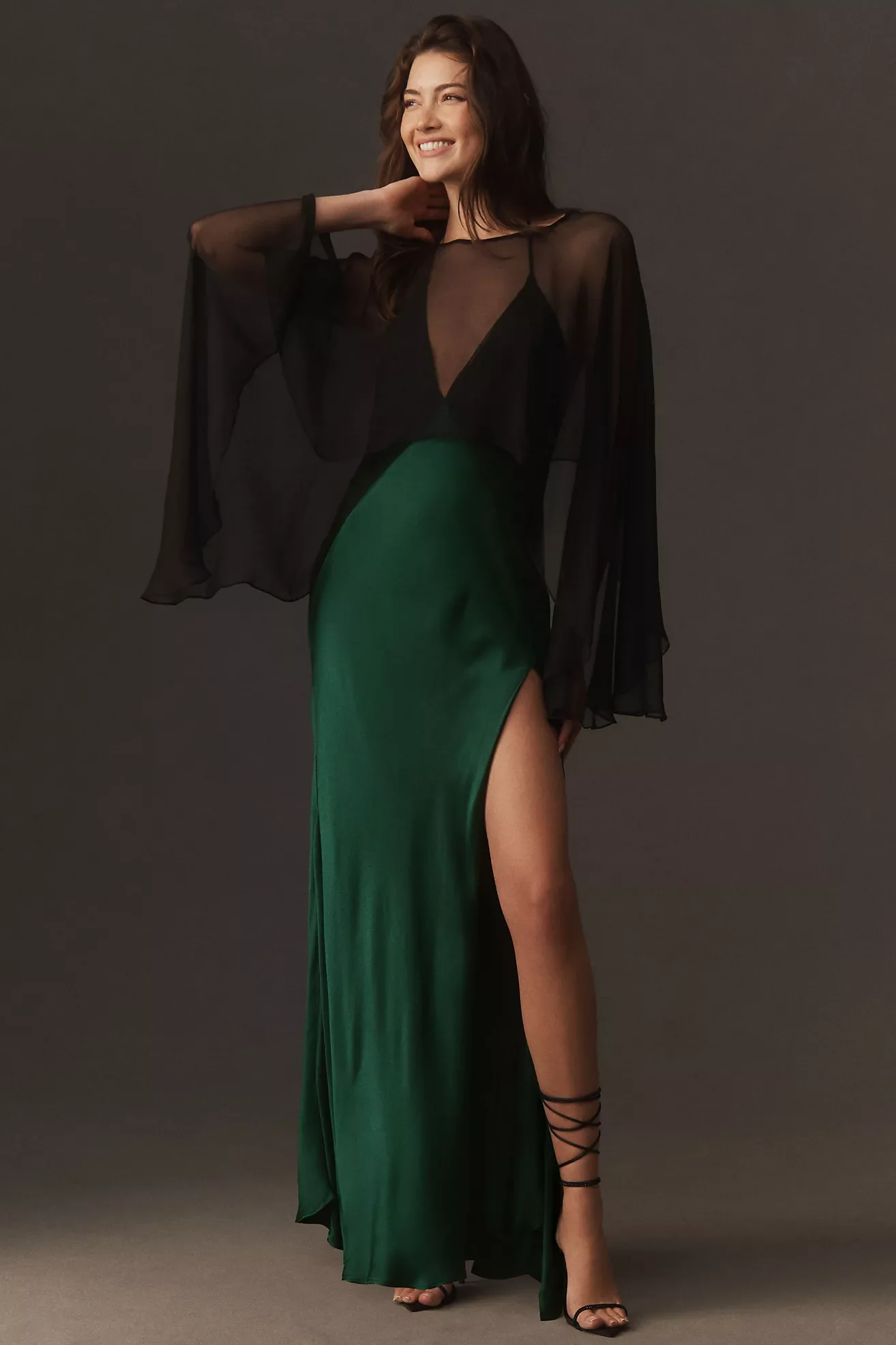 A woman with dark brown hair and a light skin tone smiling, wearing a black sheer top with a spaghetti strap dress, a high slit in a green satin skirt, and black strappy high heels, standing against a plain dark gray background.