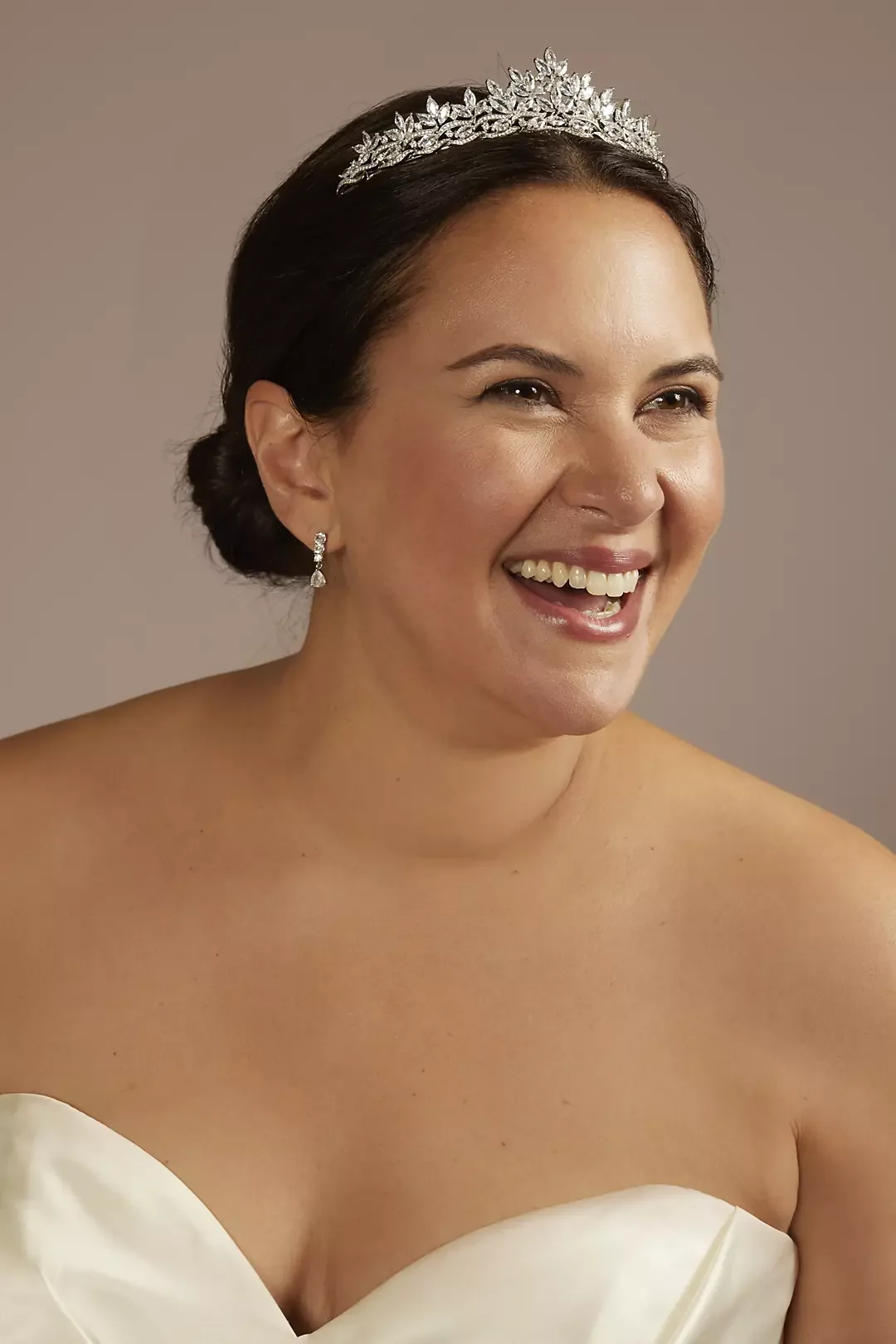 Close-up of a woman wearing a tiara, earrings, and a strapless white dress, smiling against a neutral background.