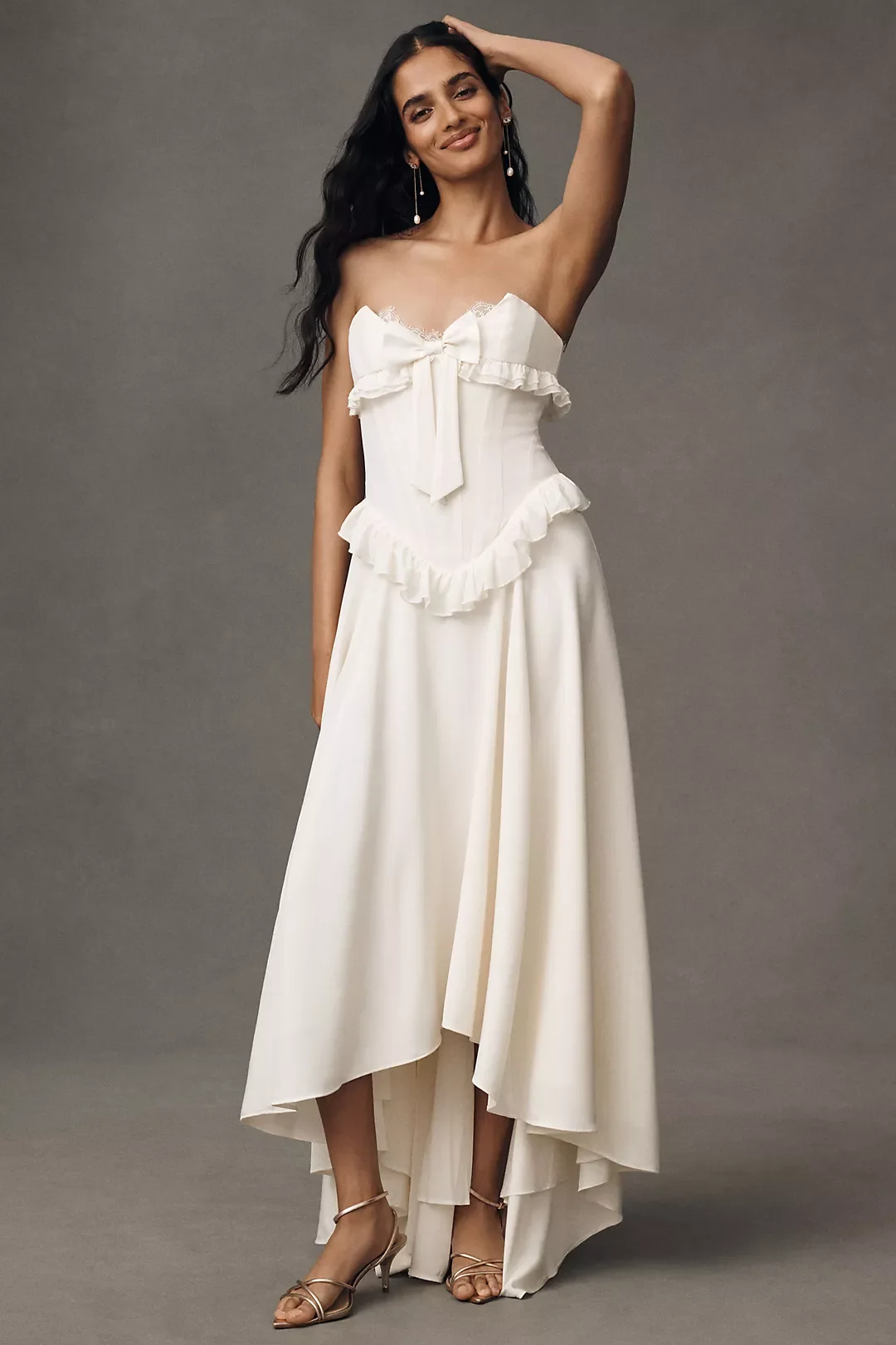 Woman in a white, ruffled, strapless dress with a bow detail and high-low hem, standing against a gray background, wearing high-heeled sandals and earrings, smiling and touching her hair.