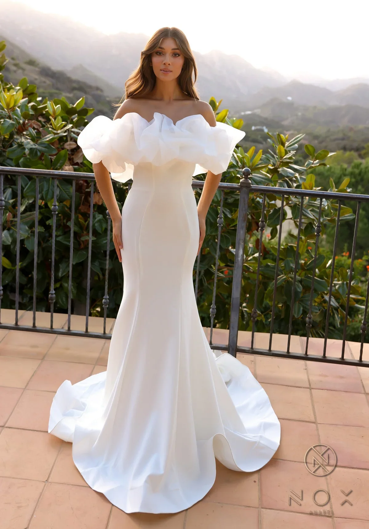 A woman in a white, off-the-shoulder, floor-length gown with ruffled sleeves stands on a tiled balcony with a mountain and green foliage background.