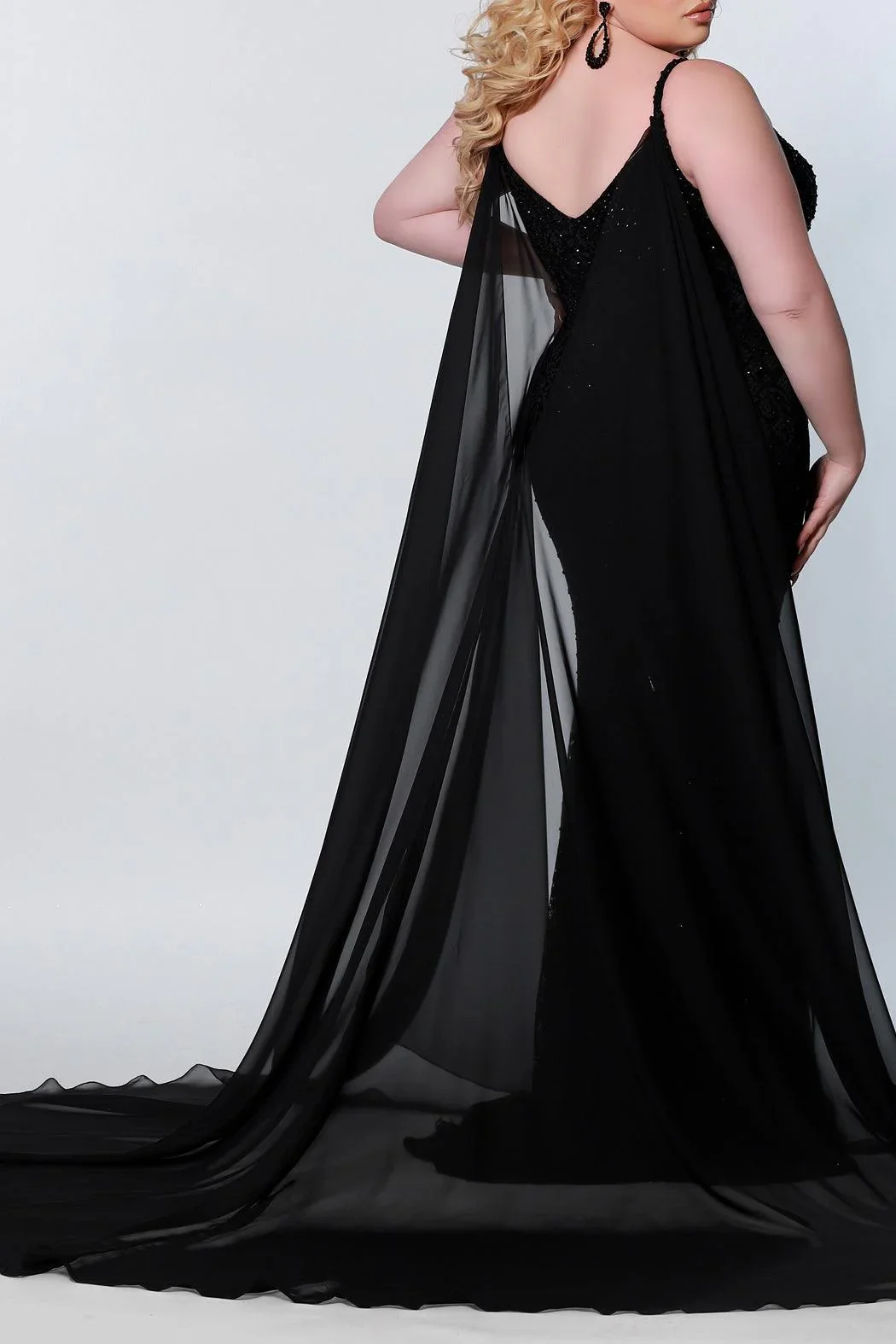 A woman wearing a black evening gown, with sheer fabric and embellishments, standing against a plain light background.