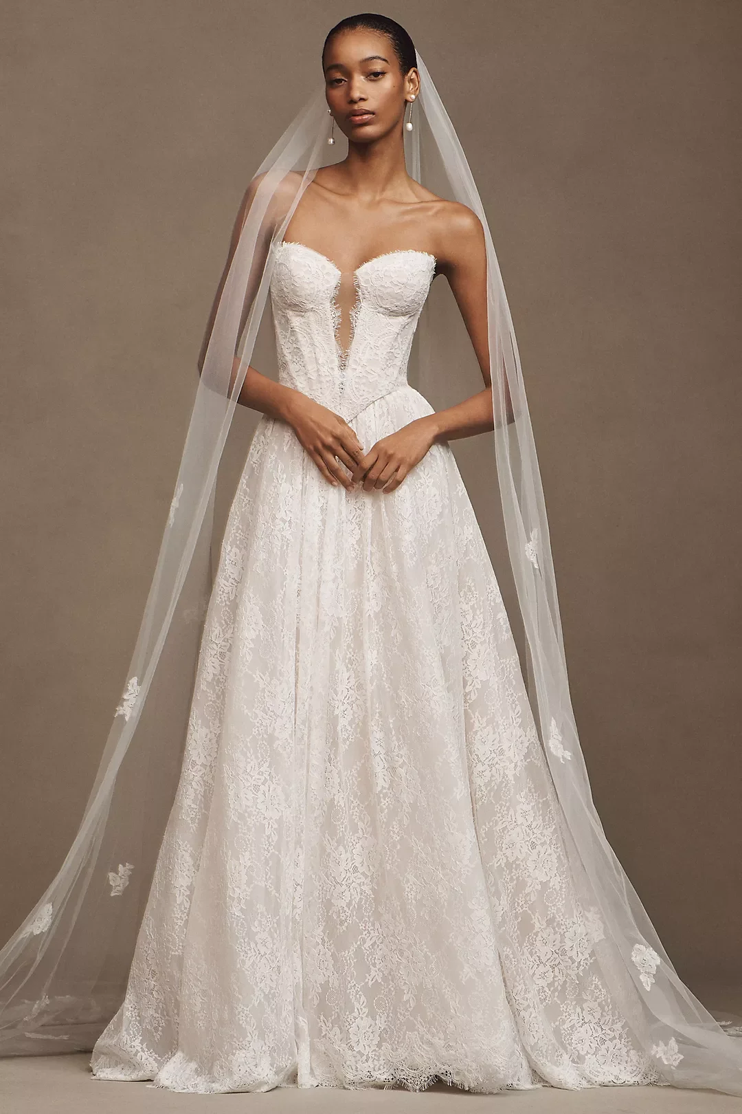 Woman in a white lace wedding dress with a sweetheart neckline, wearing a veil and pearl earrings, standing against a neutral background.