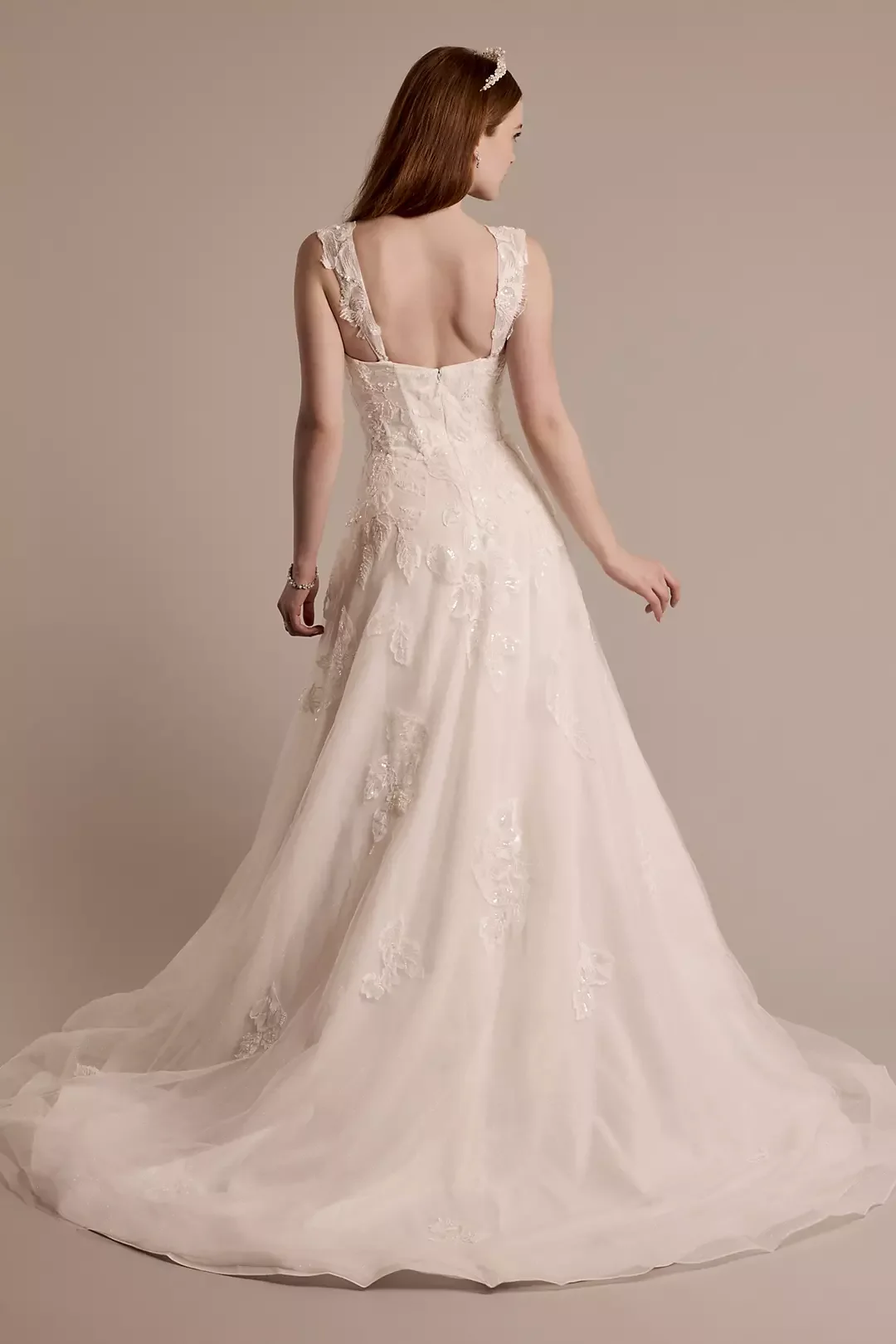 A woman wearing a white wedding gown with lace details, standing against a plain background, seen from the back.
