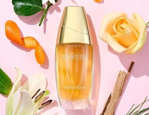 A bottle of Estée Lauder Beautiful perfume surrounded by flowers, a vanilla bean, a green leaf, an orange peel, and a pink background.