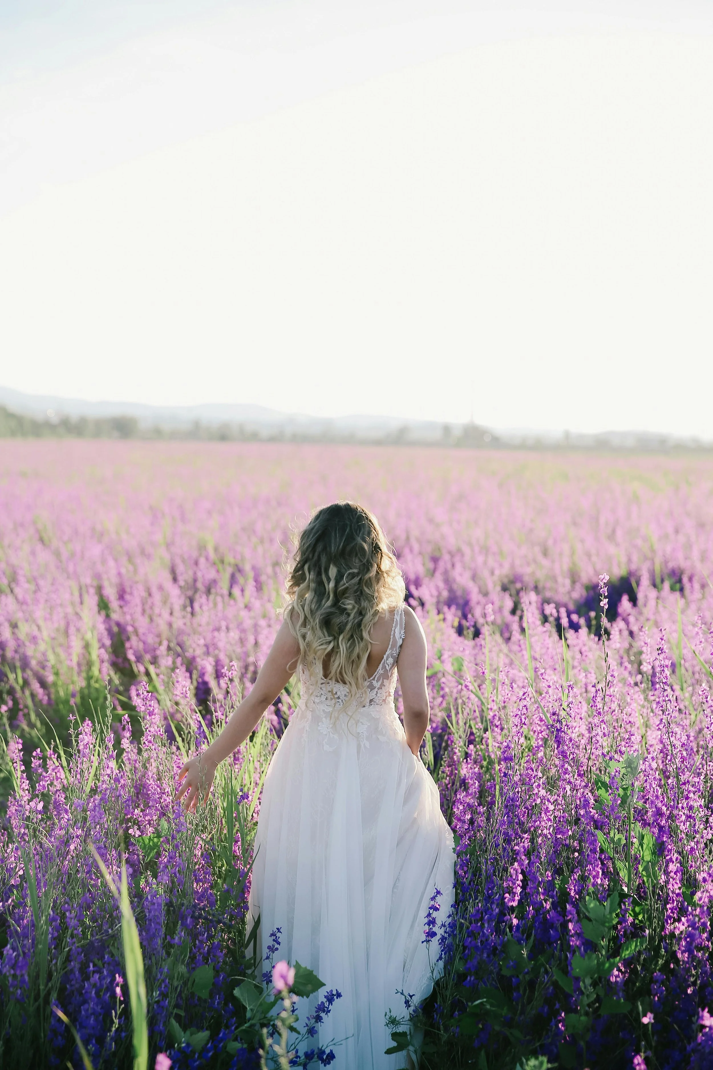 A woman in a white wedding dress walking through a field of purple lavender flowers during daylight.