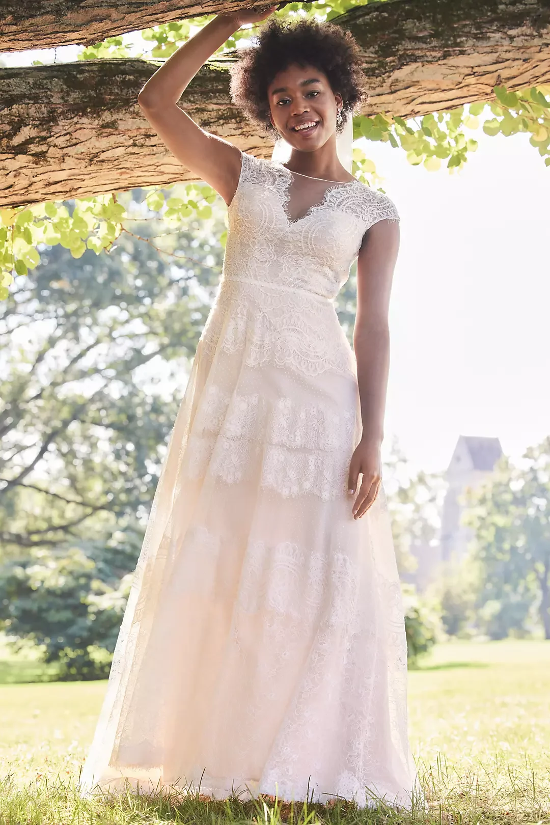 A woman wearing a white lace wedding dress standing outdoors under a large tree, smiling, with sunlight filtering through green leaves and a church visible in the background.