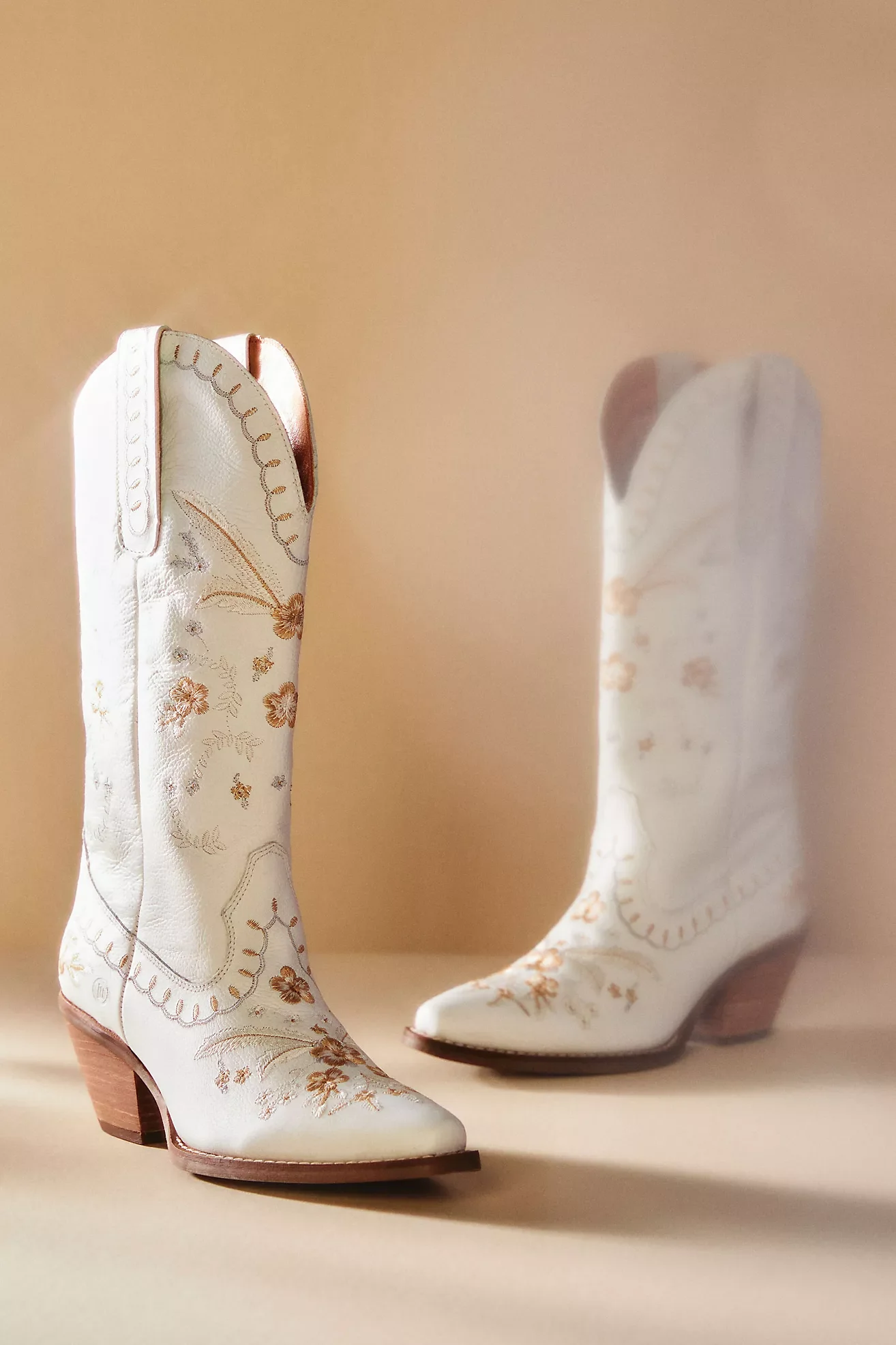 A pair of white cowboy boots with embroidery in peach and gold, featuring floral and swirl designs, with a pointed toe and stacked heel, against a plain background.