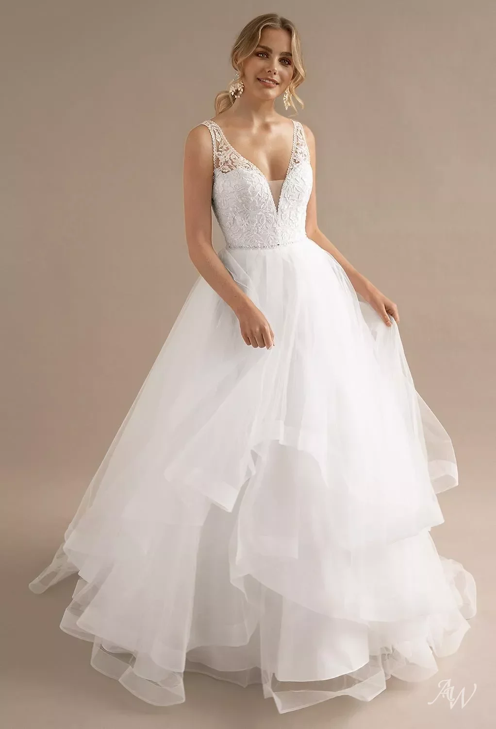 A woman in a white wedding dress with lace details on the bodice and layered tulle skirt, standing against a beige background.
