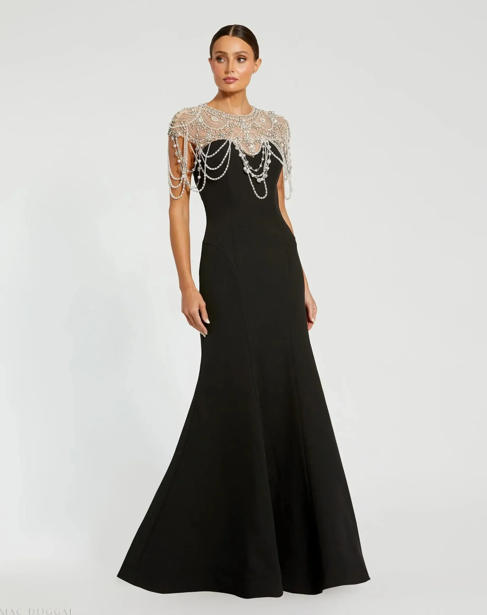 A woman in a black elegant evening gown with a fitted bodice and a long, flowing skirt, wearing a beaded cape with pearl and crystal embellishments draped over her shoulders, standing against a plain white background.