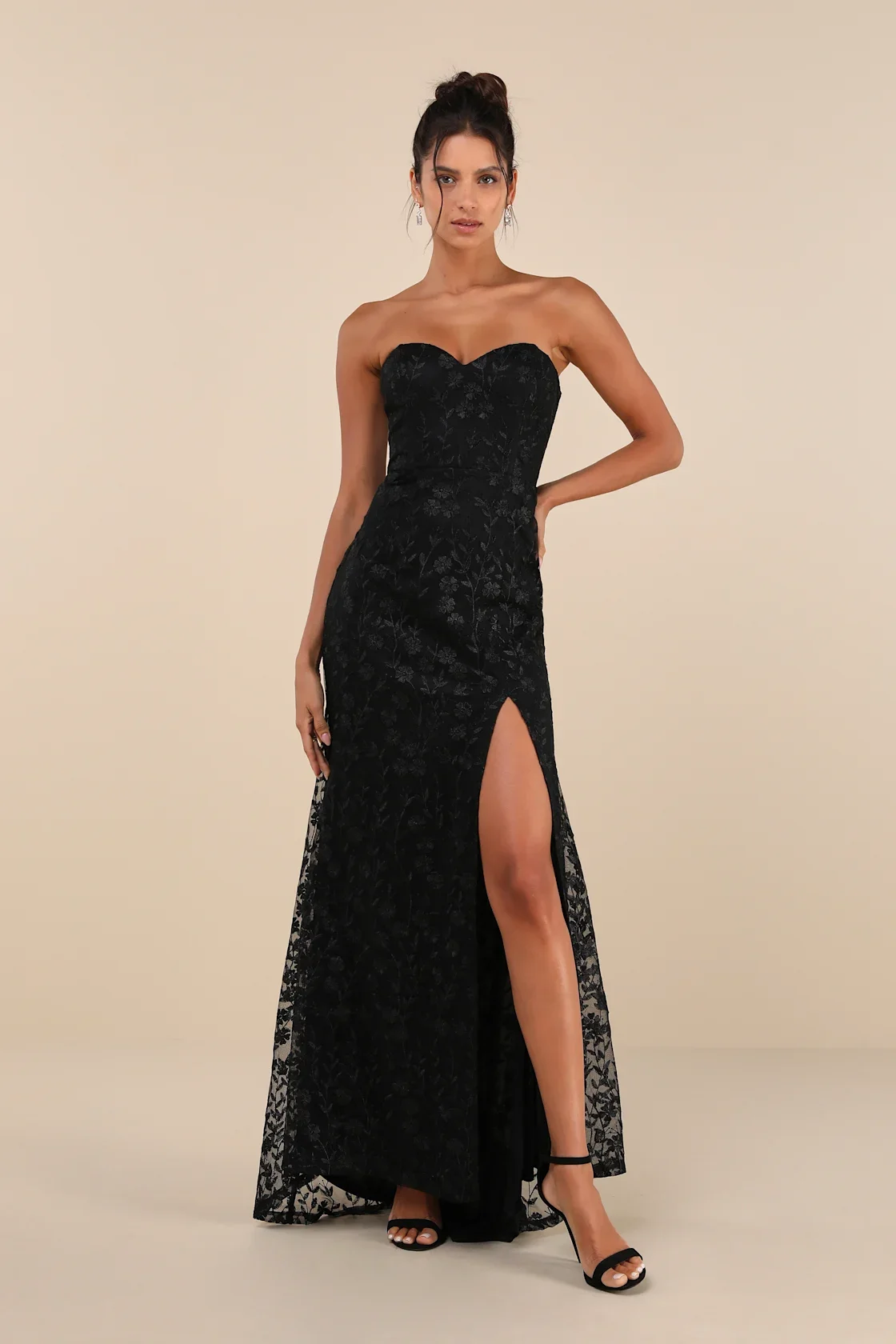 A woman in a black strapless lace gown with a high slit, black high heels, and earrings, standing against a plain beige background.