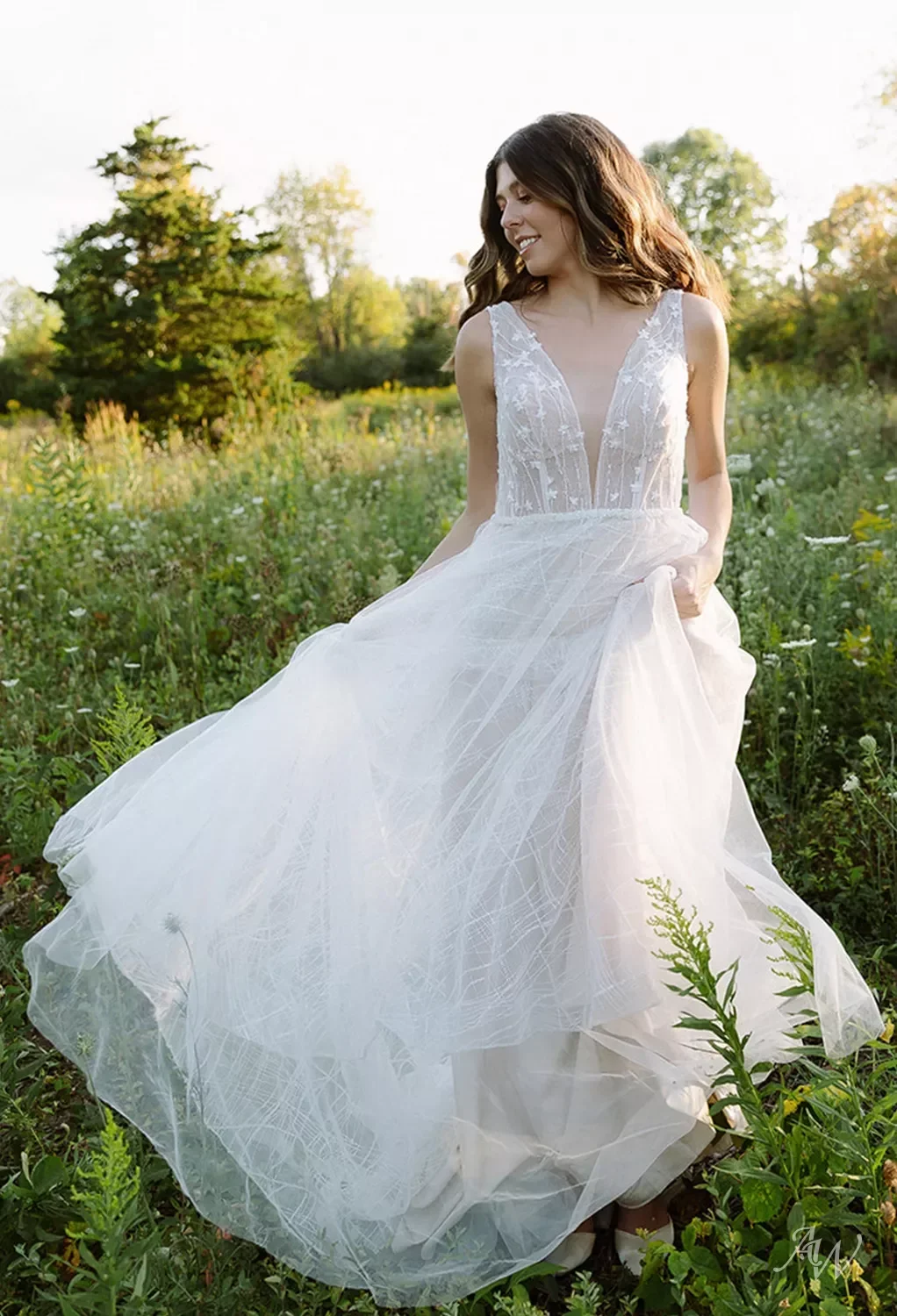 A woman in a white wedding dress standing in a green field with trees in the background during daylight.