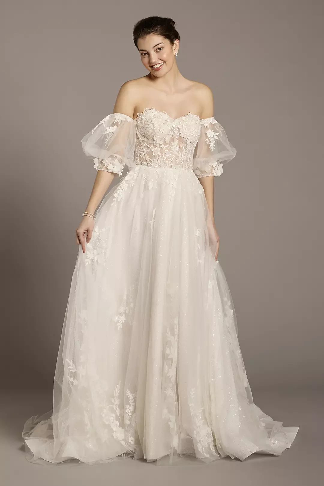 A woman wearing an off-the-shoulder wedding gown with lace and tulle details, smiling and standing against a plain gray background.