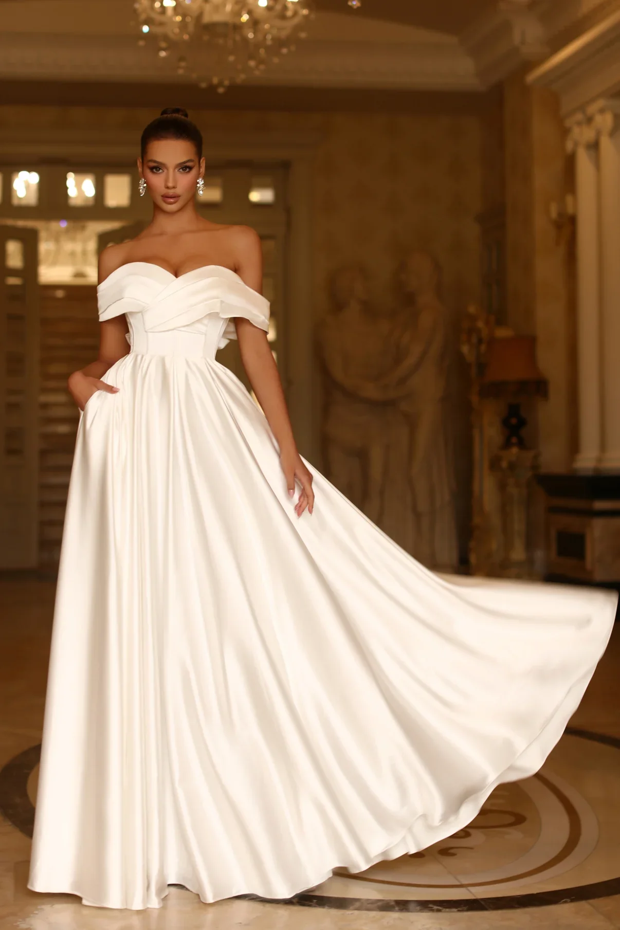 A woman in an off-the-shoulder white gown with a full skirt, posing indoors in a luxurious setting.