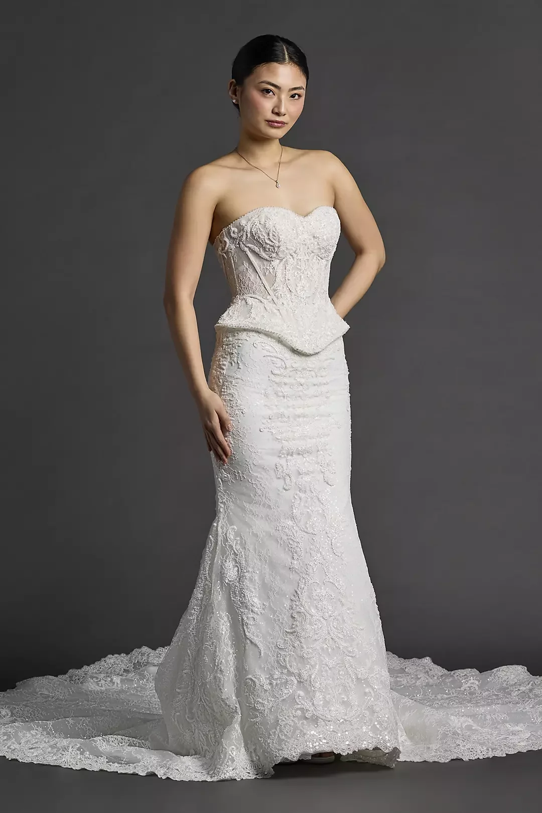 A woman in a strapless white wedding gown with intricate lace details, standing against a gray background.