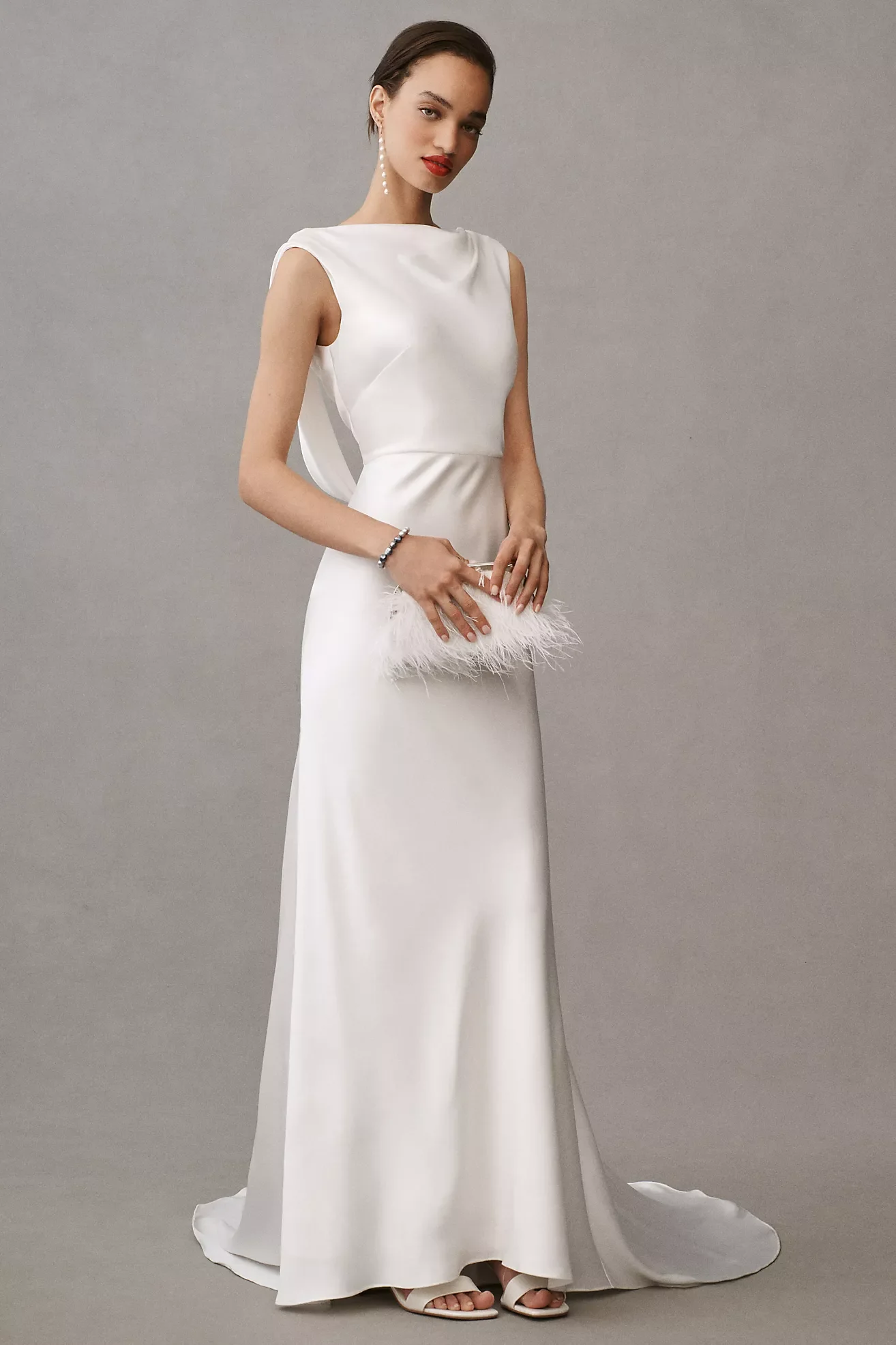 A woman wearing a sleek white satin evening gown with a feathered clutch and pearl earrings, standing against a gray background.