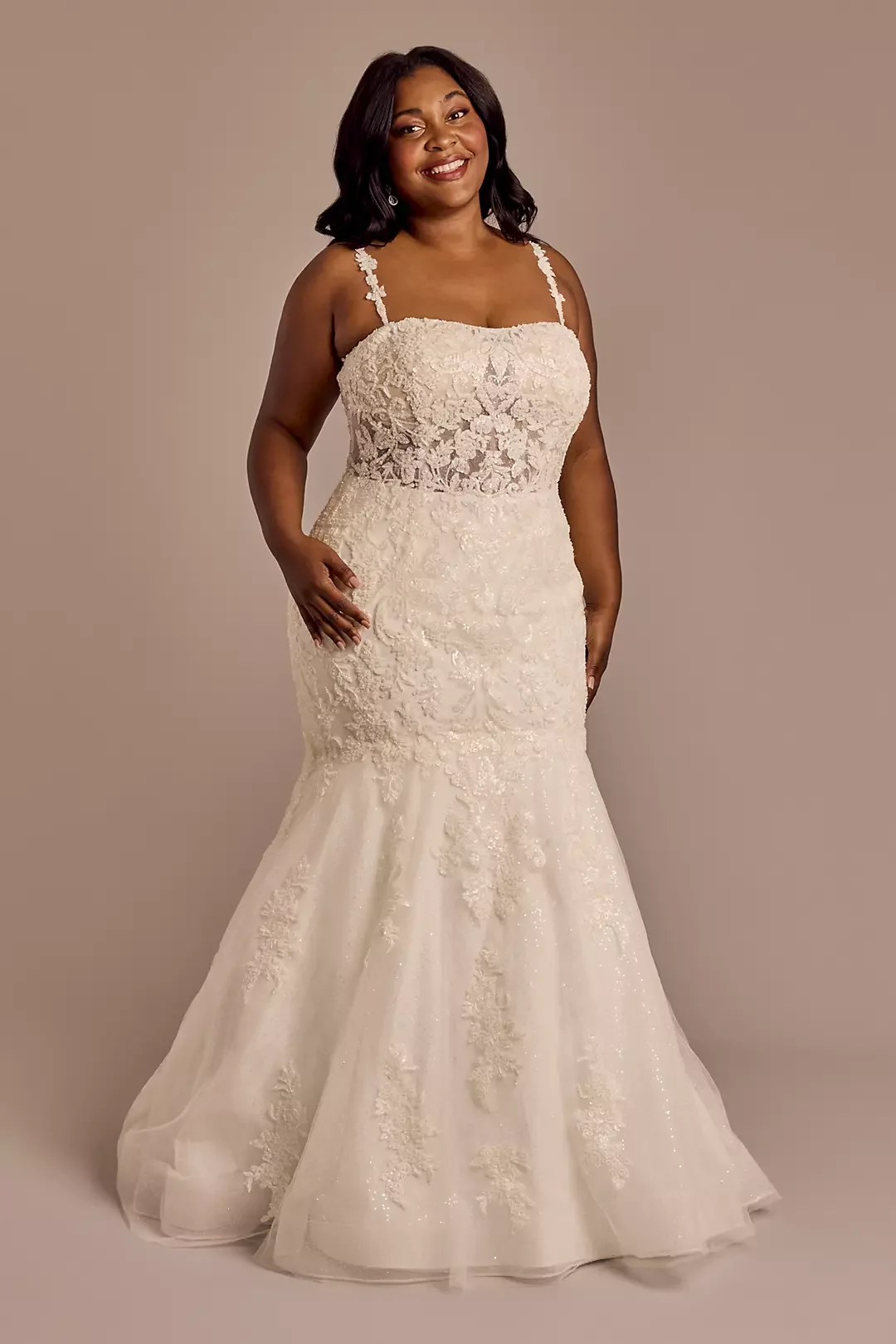 A woman in a white lace wedding gown with a fitted bodice and a flared tulle skirt, standing against a plain background, smiling with her hand resting on her hip.