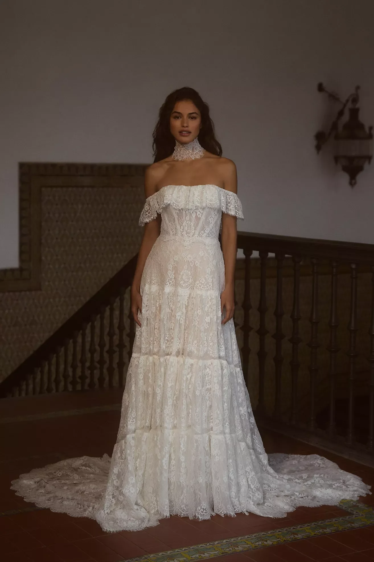 A woman is standing indoors on a staircase, wearing a white wedding dress with lace details, off-the-shoulder design, and a lace choker. The dress has a long train on the floor.