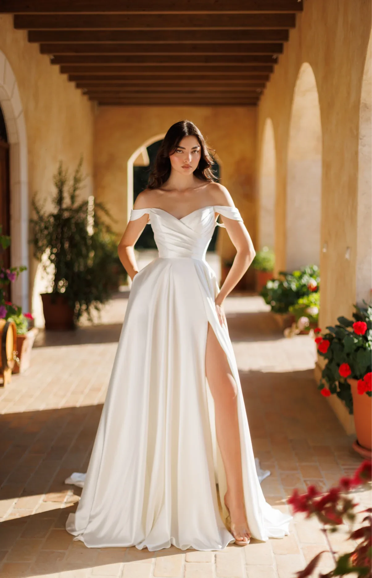 A woman in a white off-the-shoulder gown with a thigh-high slit stands in a sunlit courtyard.