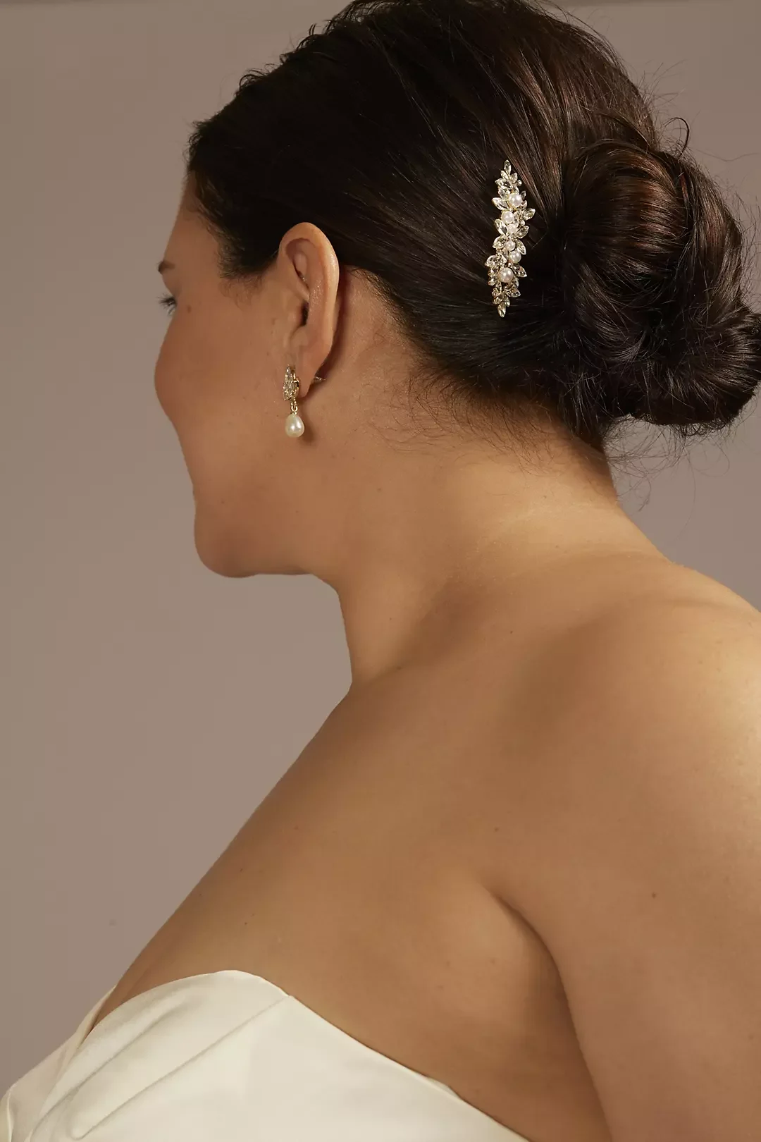 Side profile of a woman with dark hair styled in a low bun, wearing a decorative hairpin, pearl drop earrings, and a strapless white dress, against a neutral background.