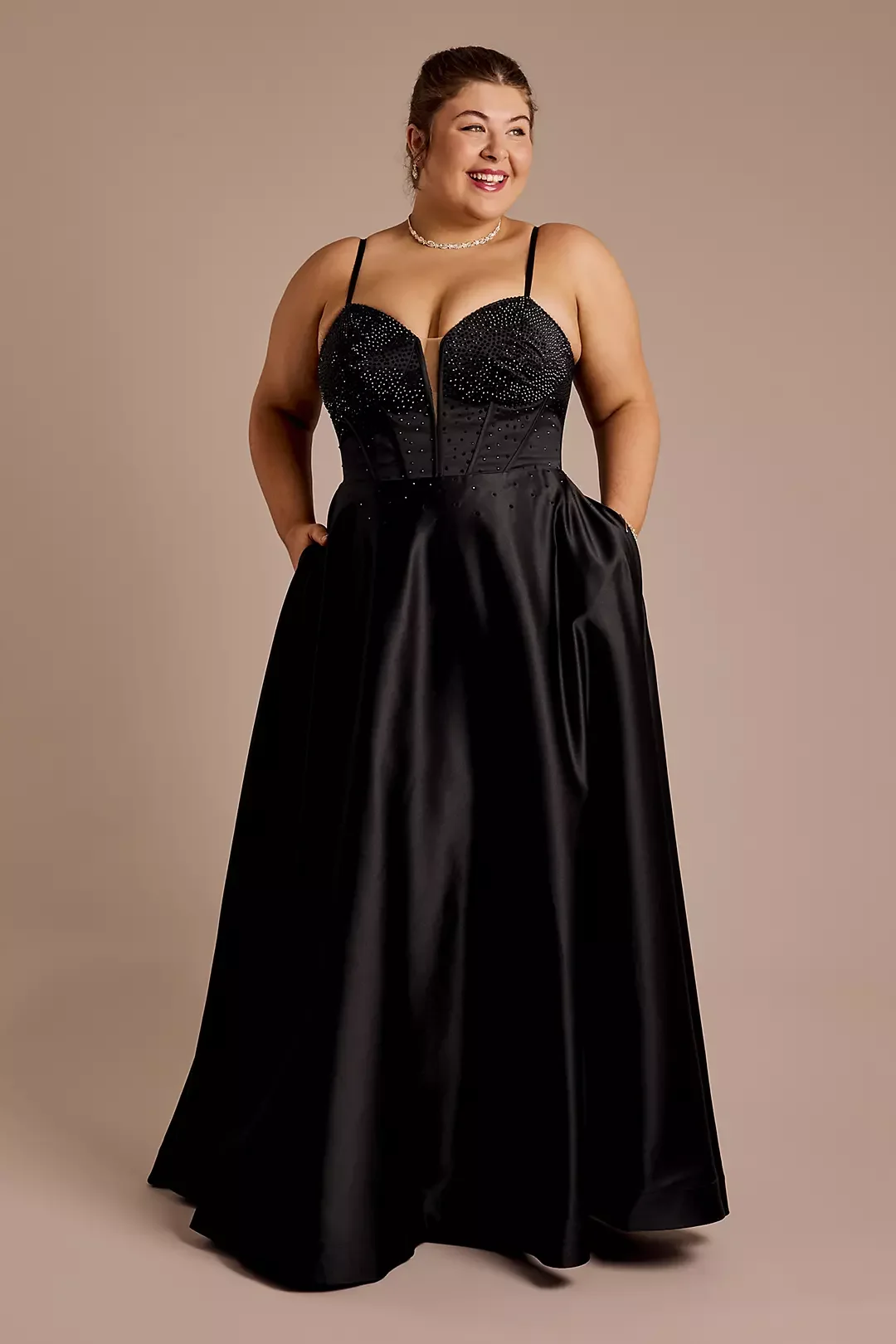 A woman in a black evening gown with a fitted, sequined bodice and a full, satin skirt, standing against a neutral background, smiling with hands in her pockets.