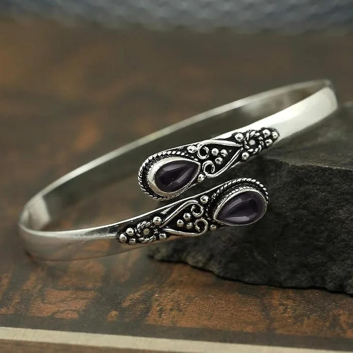 Silver bracelet with intricate design and two purple gemstones, resting on a dark stone on a wooden surface. witchy vibes aesthetic, everyday jewelry and style inspiration for women