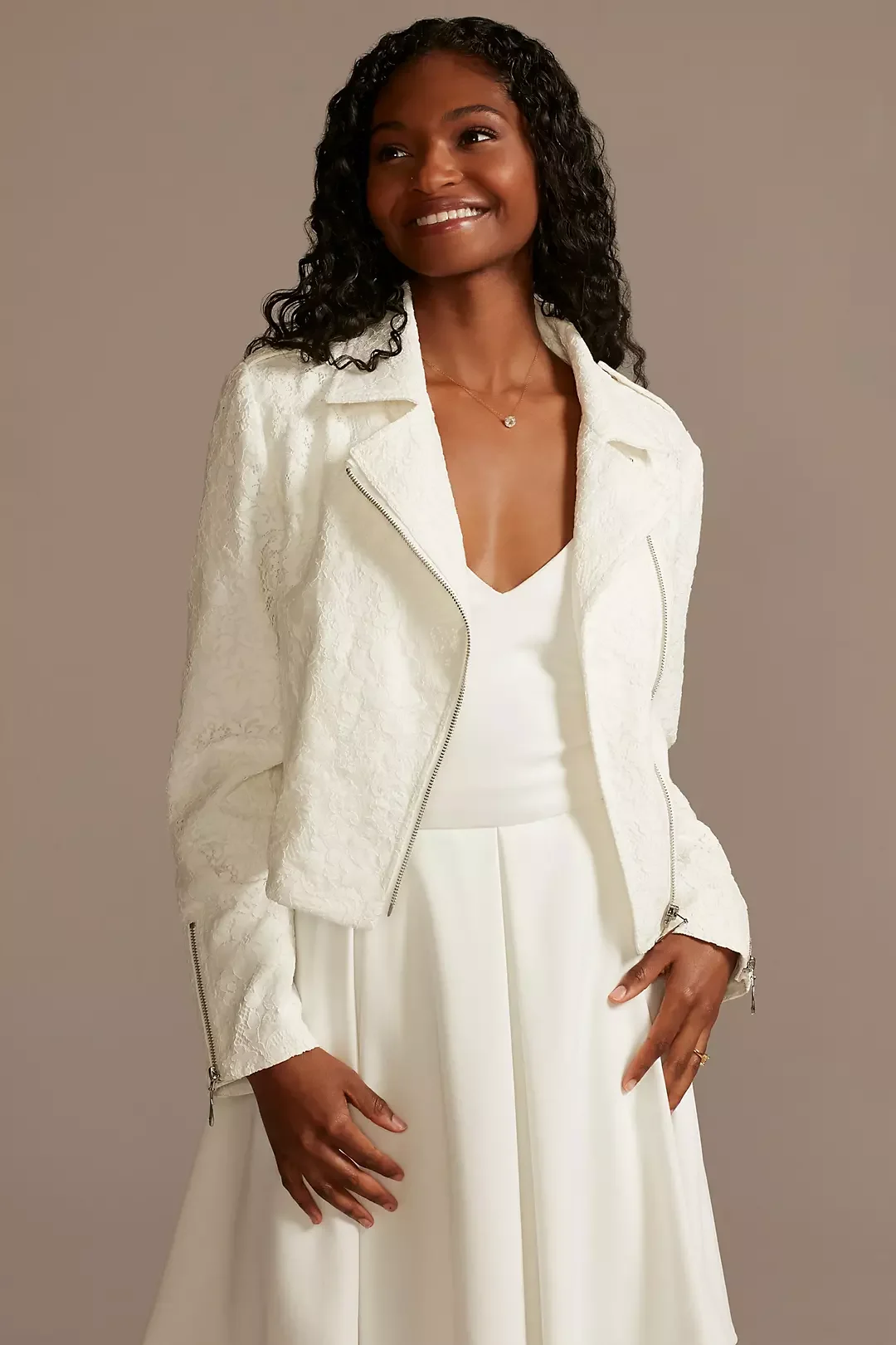 A woman with dark curly hair wearing a white textured jacket over a white dress, smiling and standing against a neutral background.