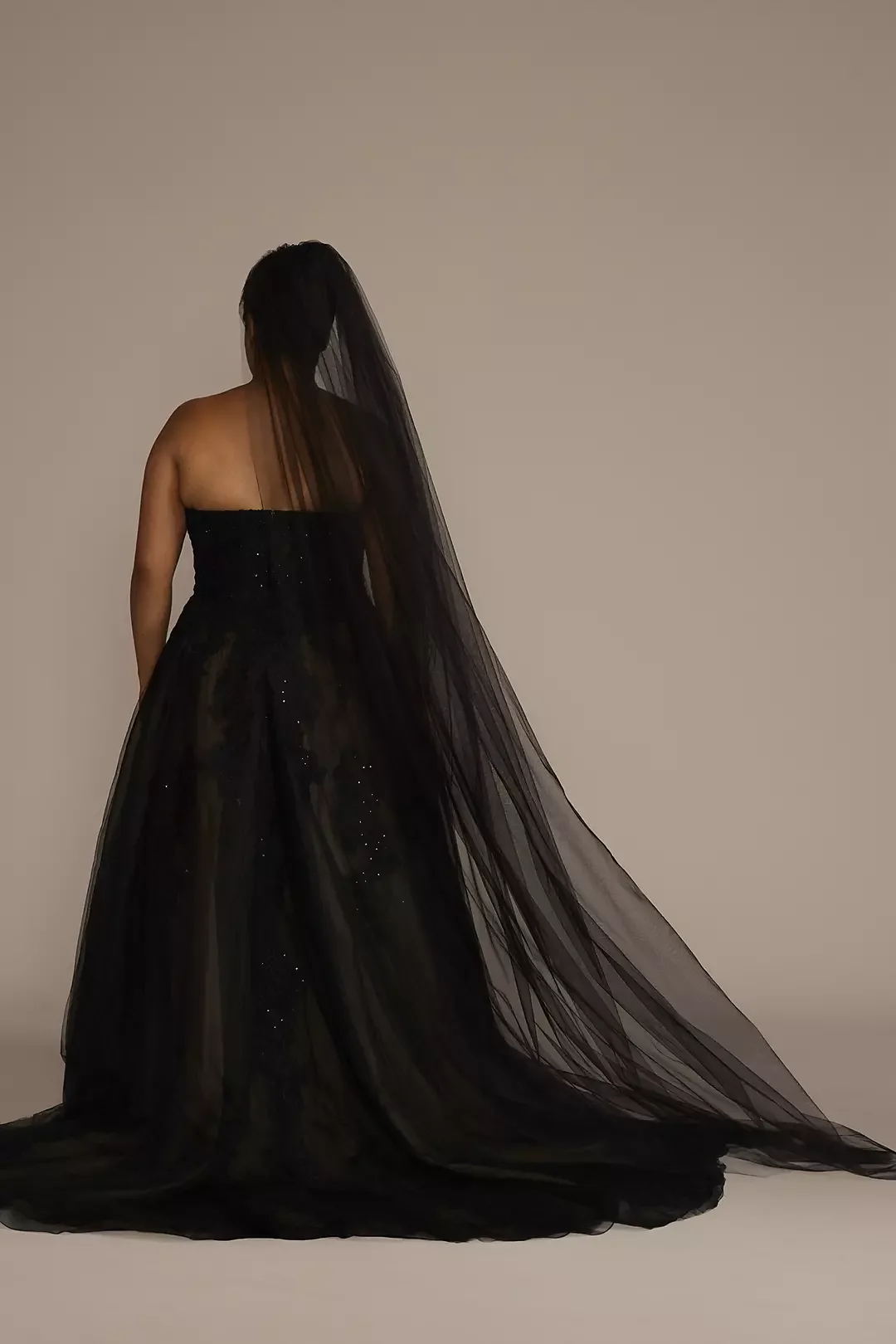 A woman in a long black gown with a sheer veil, standing with her back to the camera against a plain background.