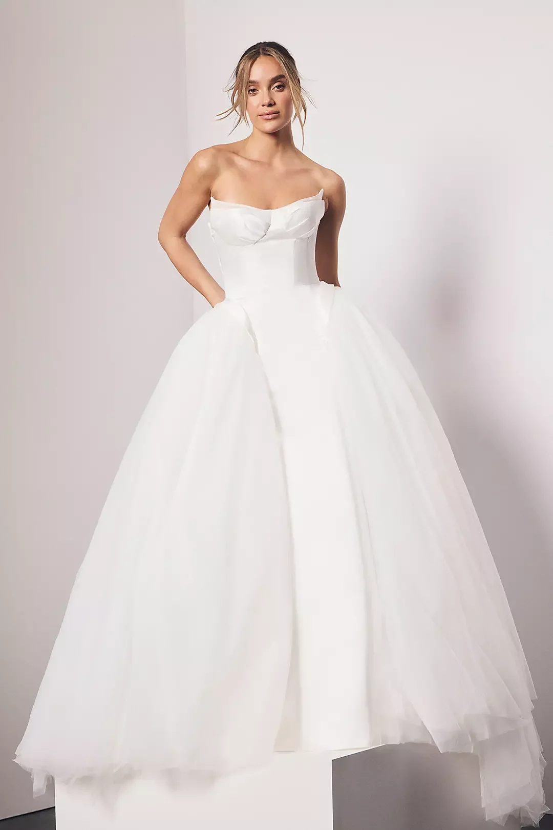 A woman in a strapless white ball gown with a fitted bodice and a full, layered skirt, posing against a neutral background.