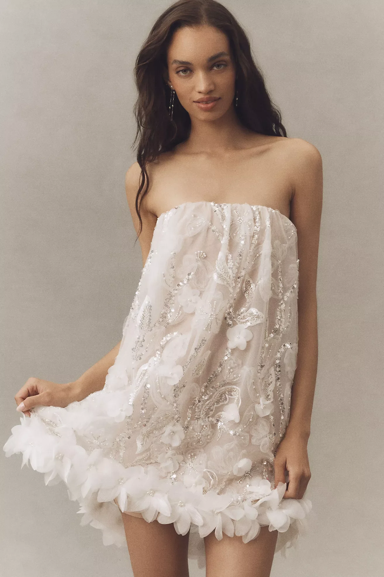 Young woman with dark wavy hair wearing a strapless, embellished white dress with floral details, standing against a plain gray wall.