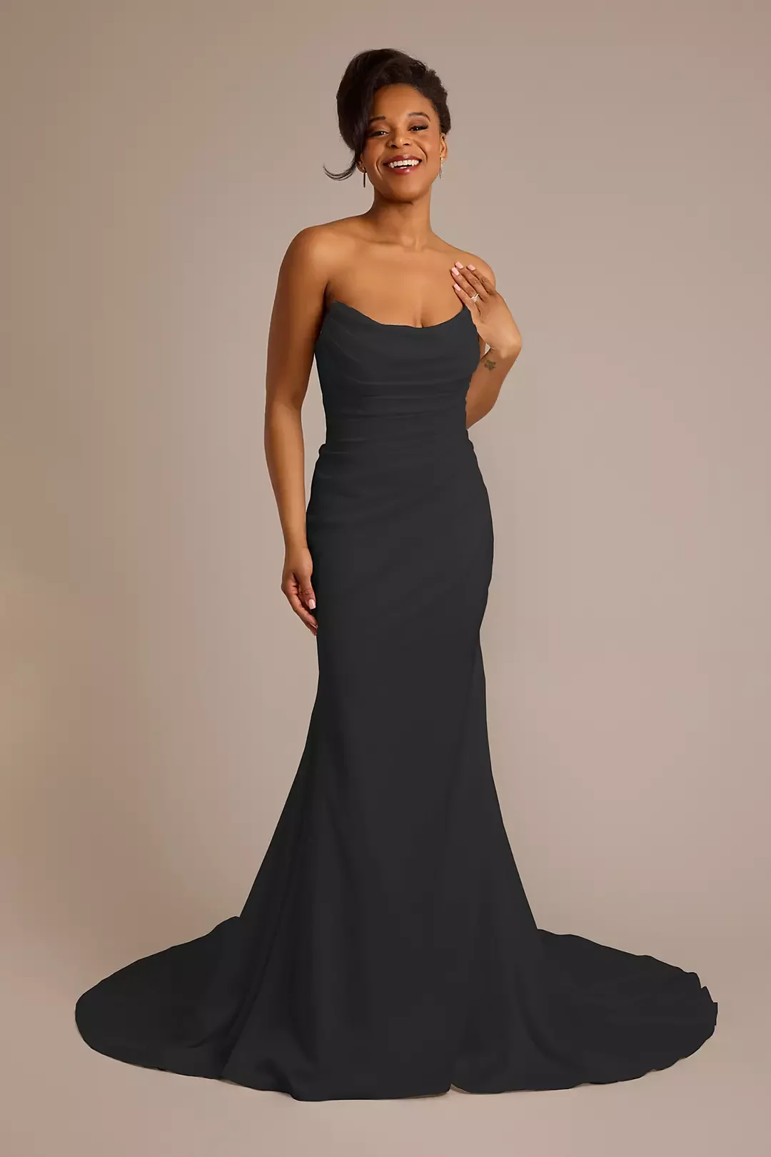 Woman in a strapless black evening gown, smiling, standing against a plain beige background.