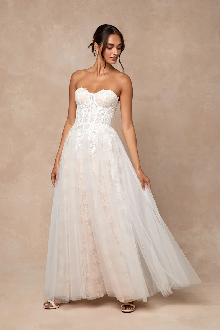 Woman in a strapless white wedding dress with lace details, standing against a beige background.