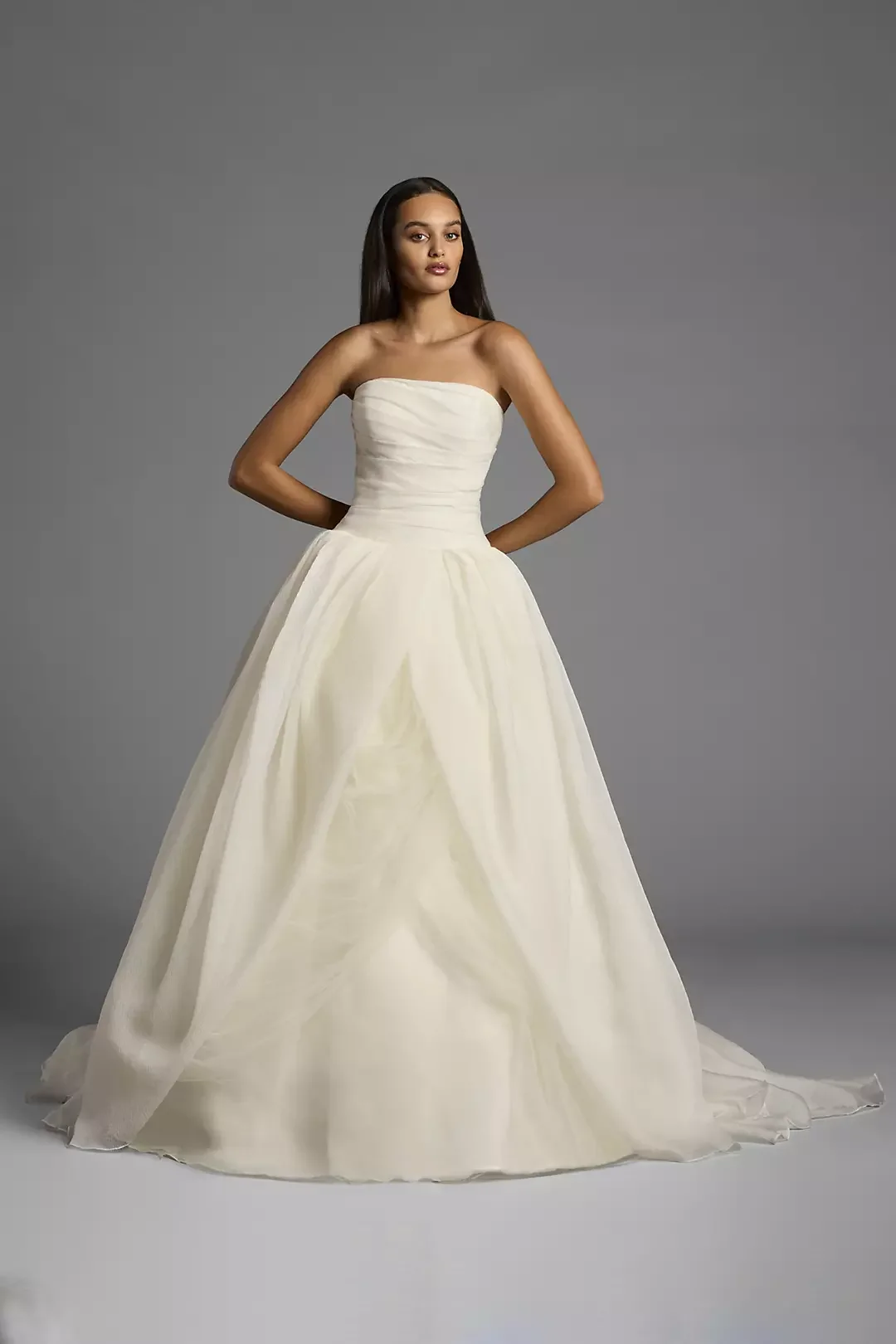 A woman wearing a wedding dress with a strapless bodice and a full, flowing skirt stands against a gray background.