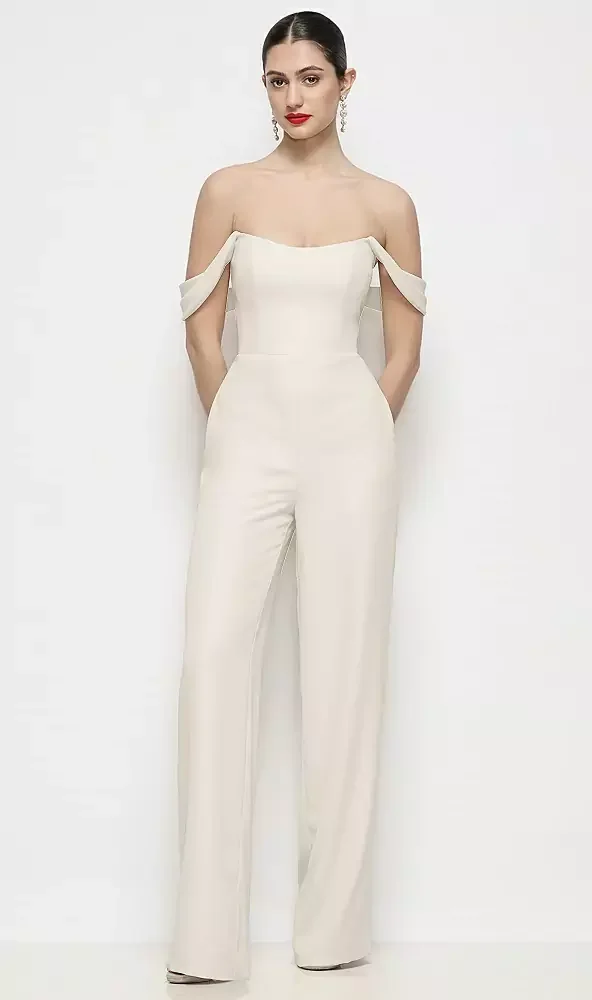 A woman stands against a white background wearing an off-the-shoulder cream-colored jumpsuit with wide legs and draped shoulder details.