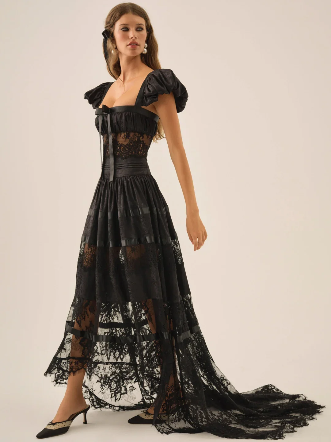 A woman in a black lace and satin evening gown with puffed sleeves and a train, standing against a plain light-colored background.