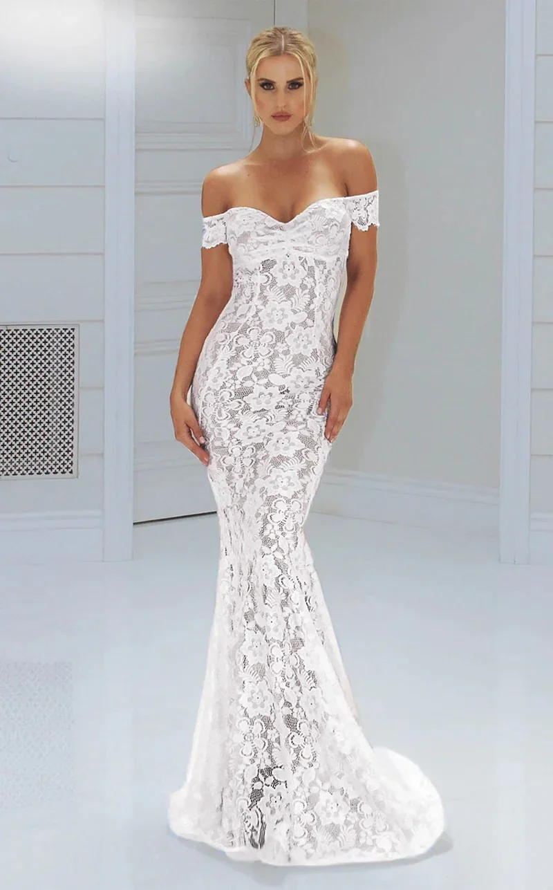 Woman wearing a white off-the-shoulder lace wedding dress with floral pattern, standing in a white room.