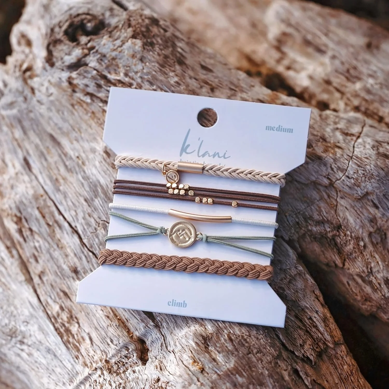 Six bracelets displayed on a white card, laid on a weathered wood surface. The bracelets vary in colors from beige, brown, white, to green, and include braided, cord, and metal designs.