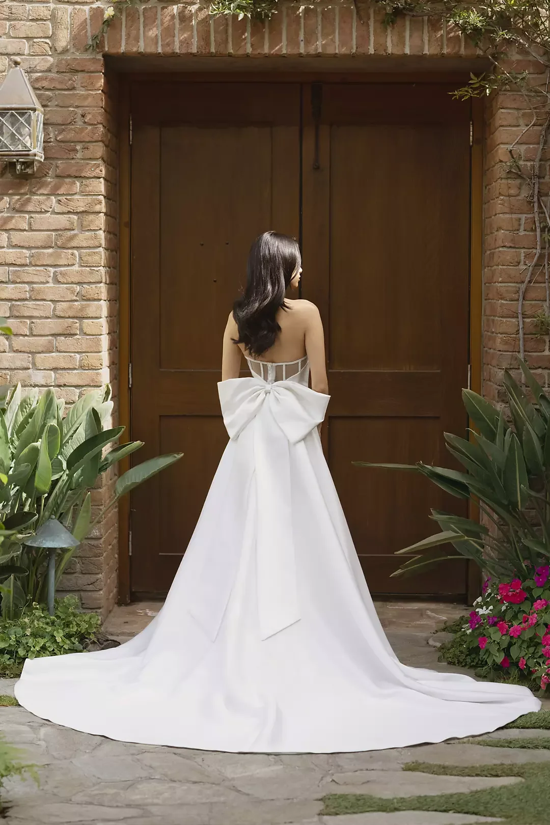 Woman standing in front of a wooden door, wearing a white wedding dress with a large bow at the back, surrounded by green plants and flowers.