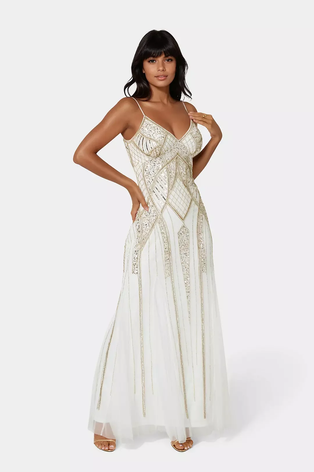 Woman in a white, gold-embellished, sleeveless dress with spaghetti straps, standing against a plain light background.