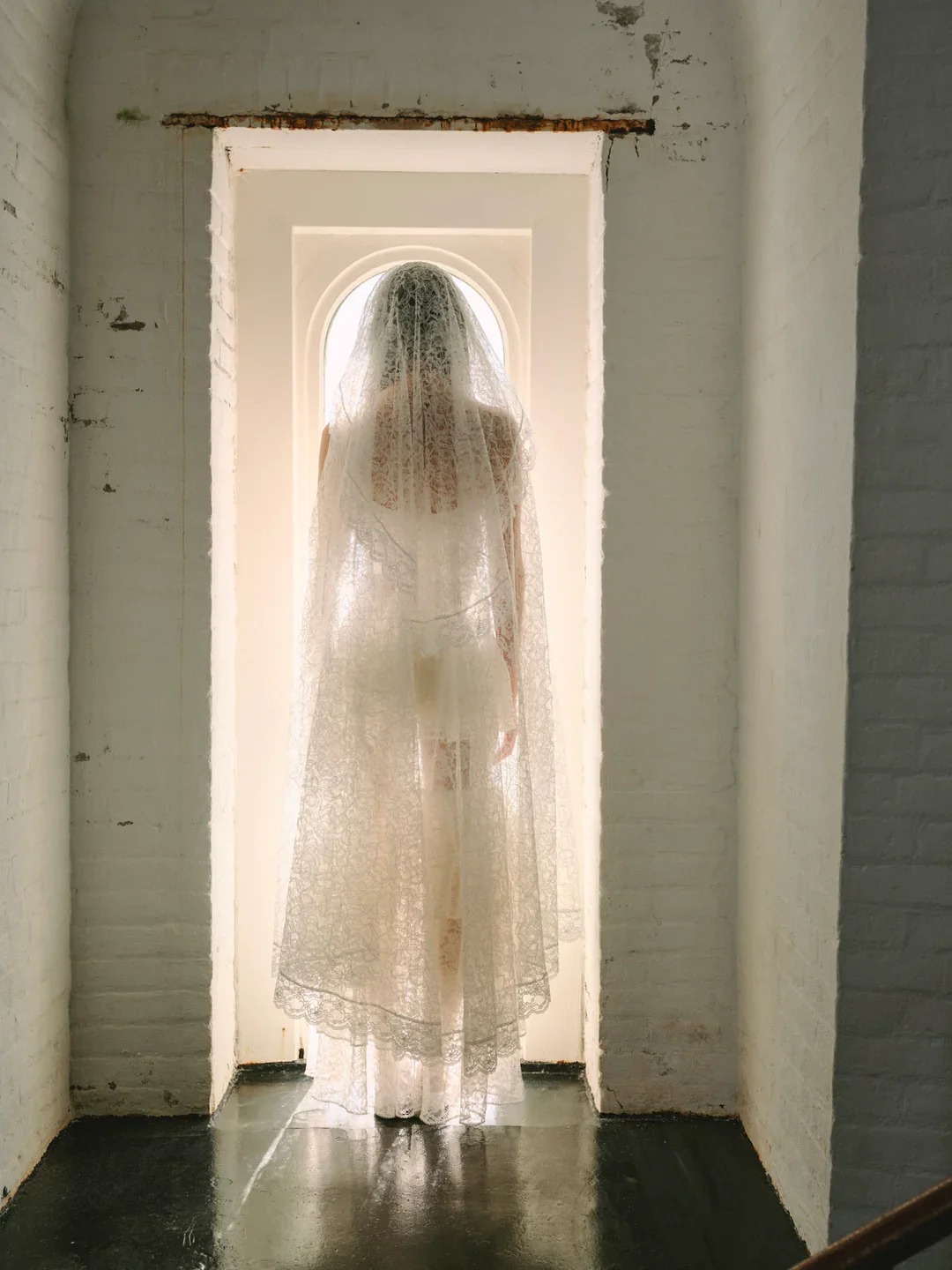 A woman dressed in a lace wedding gown and veil standing in front of a small arched window, seen from behind, in a white brick room.