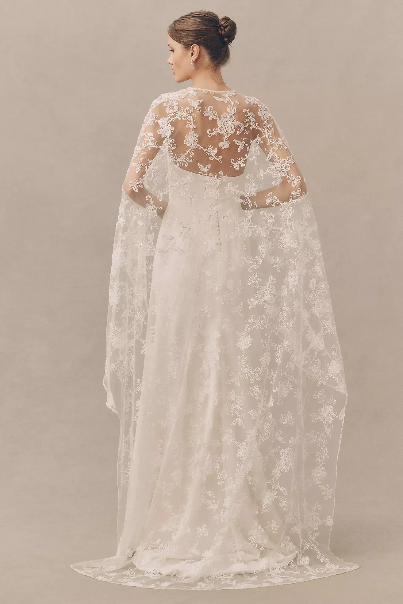 A woman in a white wedding dress with intricate embroidery, standing against a neutral background, facing sideways.